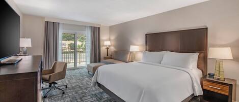 Una habitación de hotel con una cama grande, un escritorio con computadora, una silla y una mesita de noche con lámpara.
