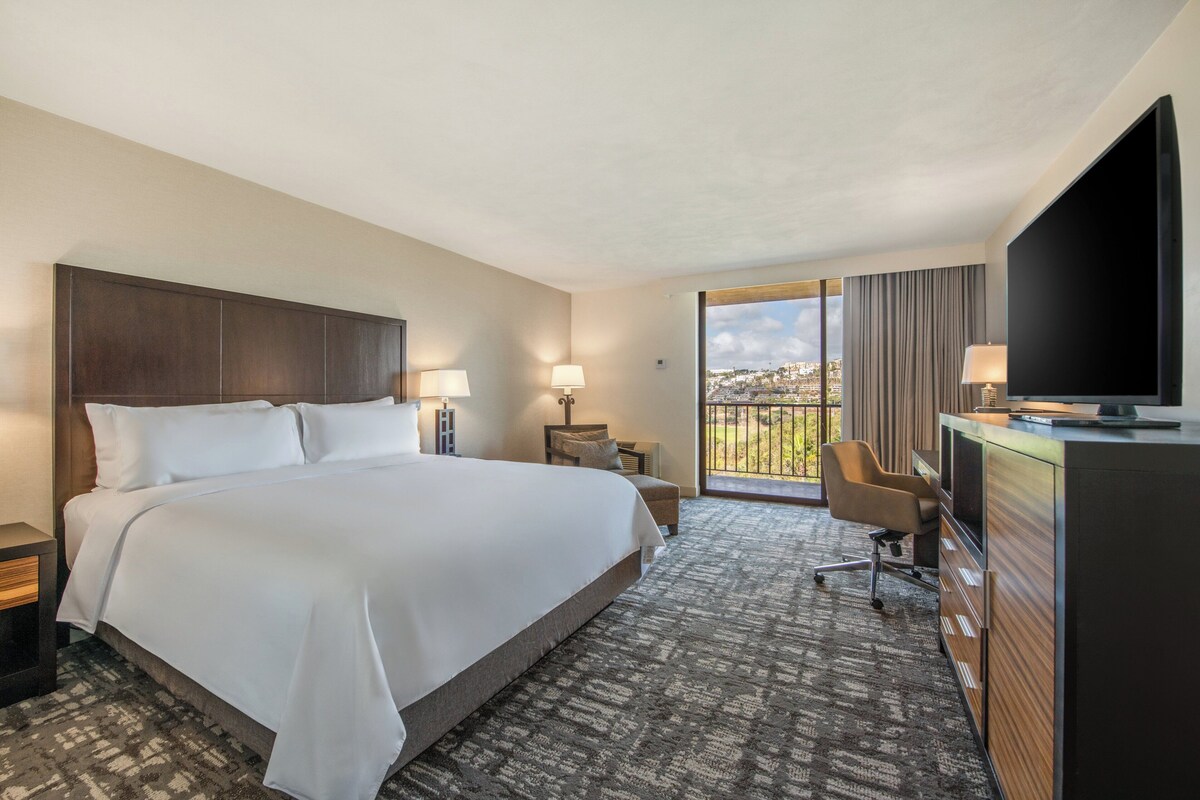 Een hotelkamer met een groot bed, een bureau, een televisie en een balkon met uitzicht over de stad.