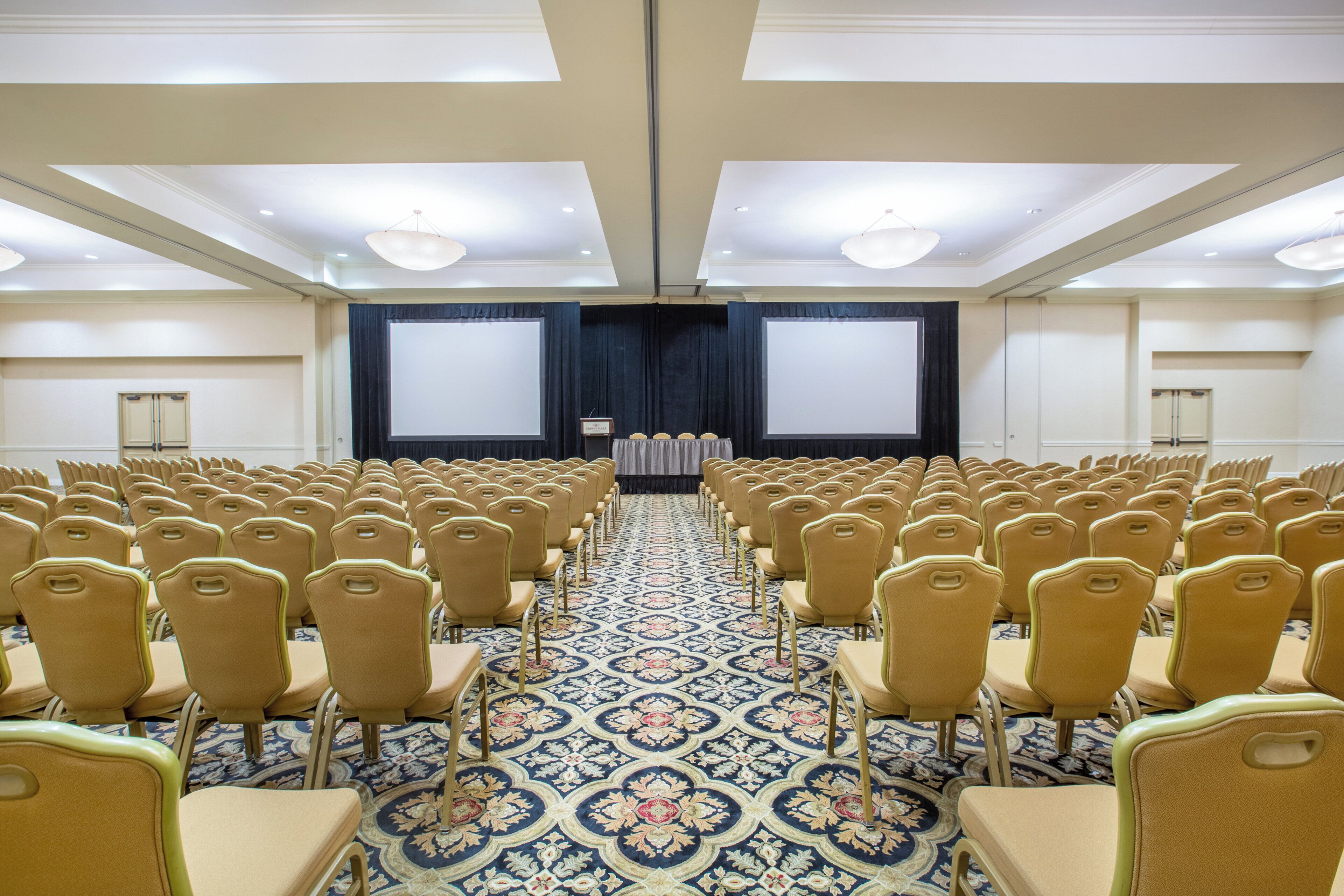 A conference room with rows of chairs facing a stage with a screen, a patterned carpet, and a ceiling with recessed lighting.