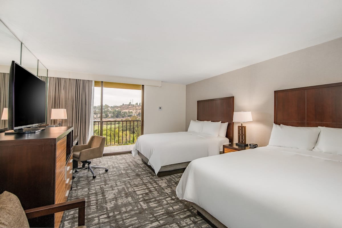 A hotel room with two beds, a desk with a TV, and a view of a cityscape through large windows.