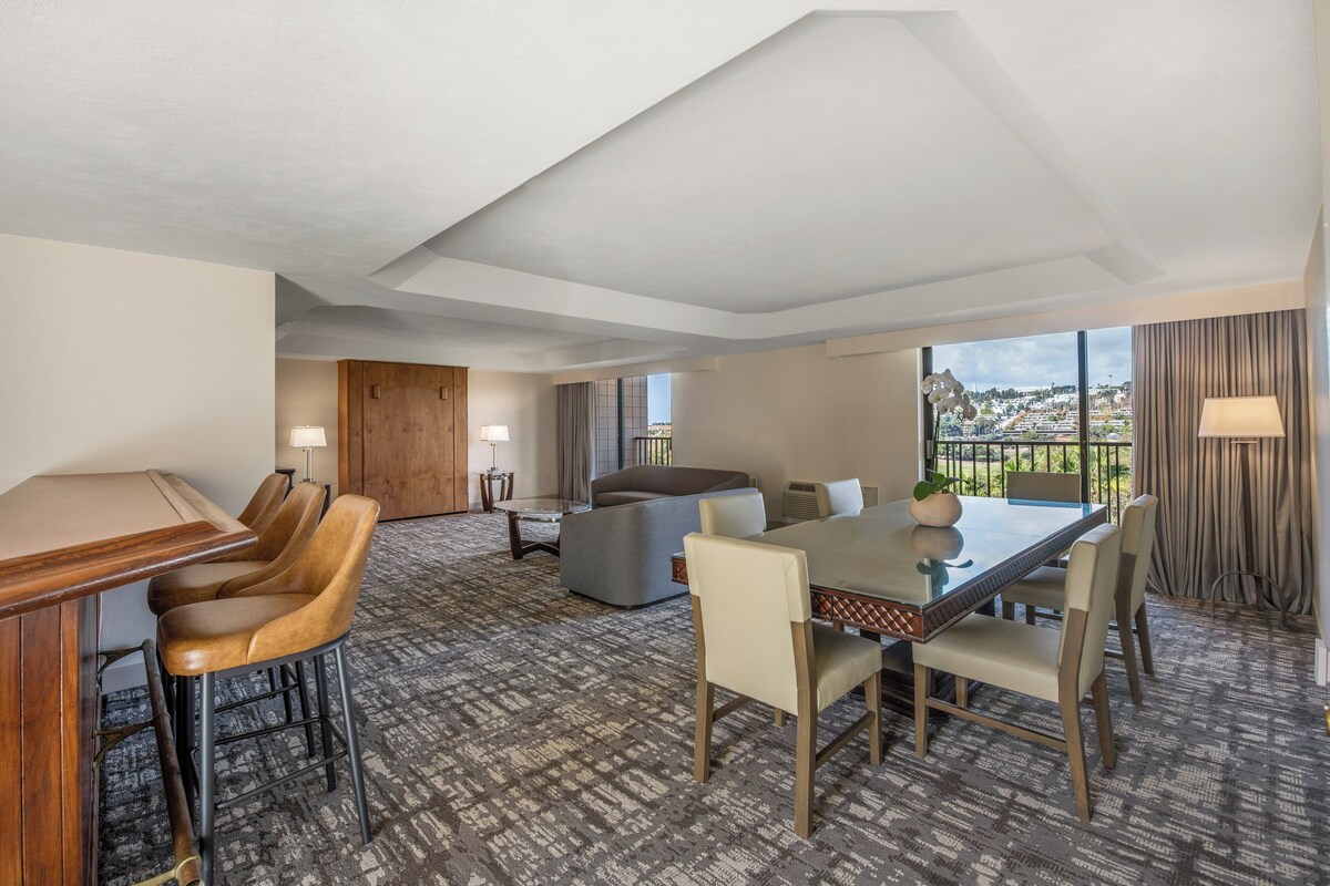 Hotelkamer met een eethoek, een bank, een groot raam met uitzicht over de stad en een houten deur.
