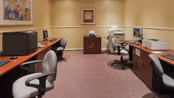 A conference room with four desks, each equipped with a computer, keyboard, and mouse. There are two printers on the right side, and a filing cabinet in the center. The room has a carpeted floor and beige walls.
