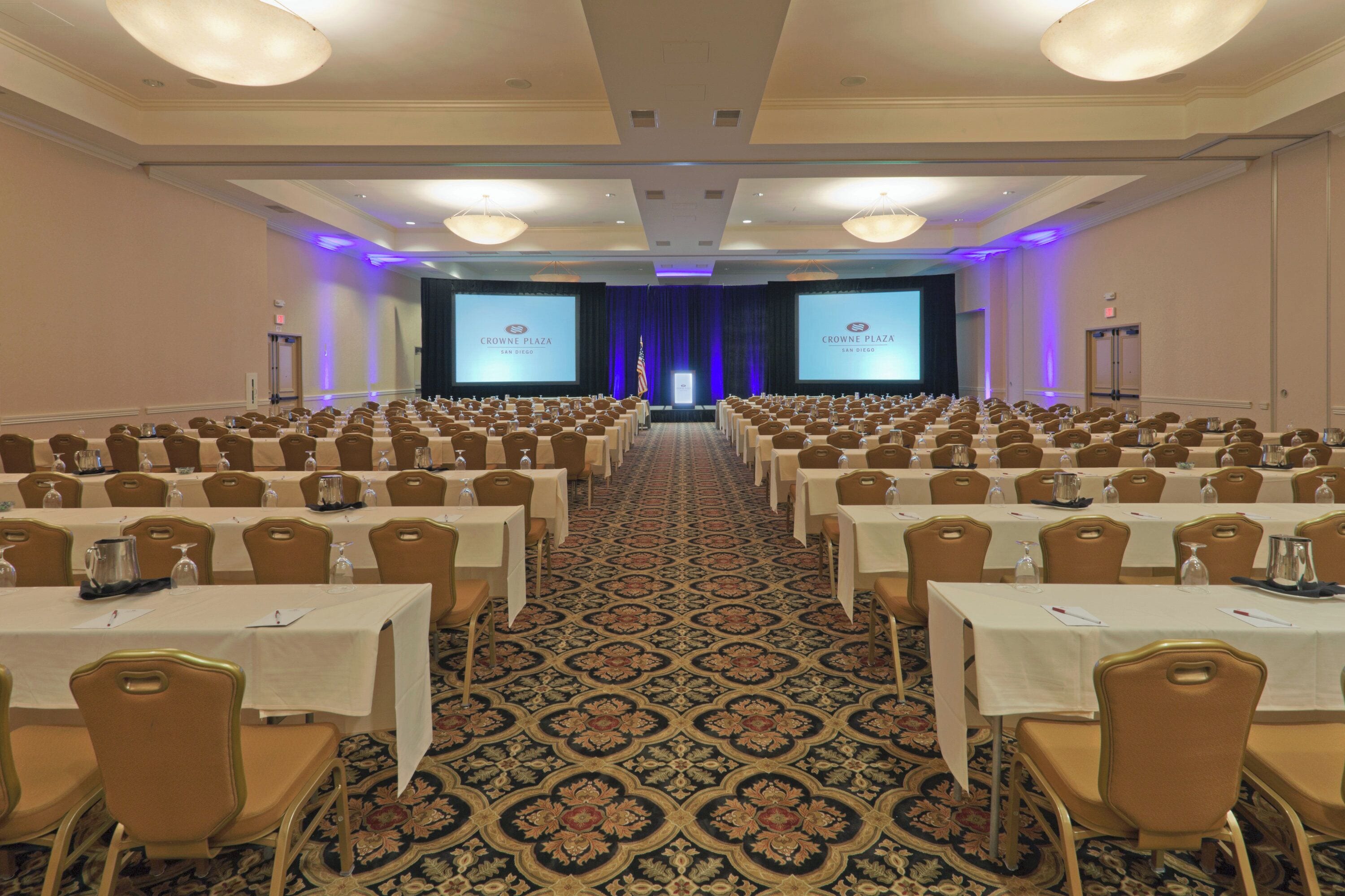 A conference room with rows of tables and chairs, a carpeted floor, and a stage with a screen displaying 'Crowne Plaza San Diego'.