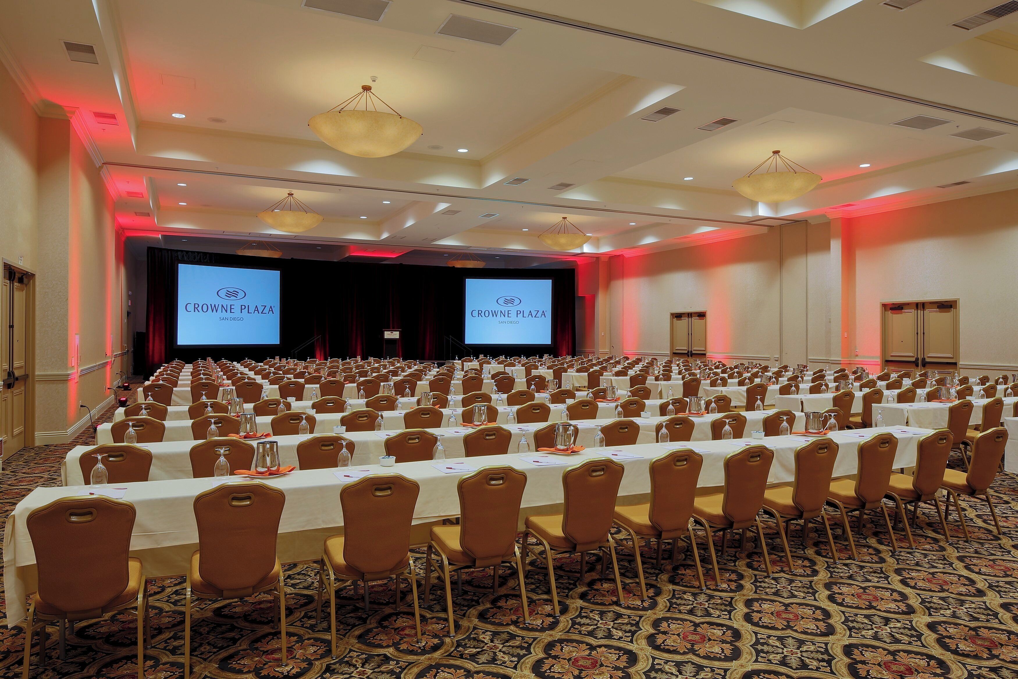 A conference room with rows of tables and chairs, a stage with a screen, and a patterned carpet.