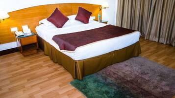 Habitación de hotel con cabecera de madera, cama con sábanas blancas y dos almohadas rojas, mesita de noche con lámpara y alfombra verde.