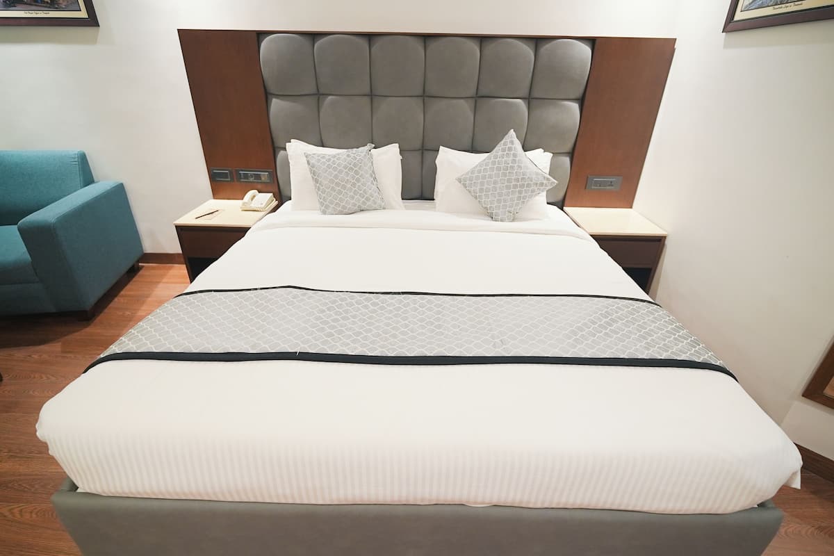 A hotel room with a large bed, a tufted headboard, two bedside tables with lamps, a blue armchair, and a framed picture on the wall.