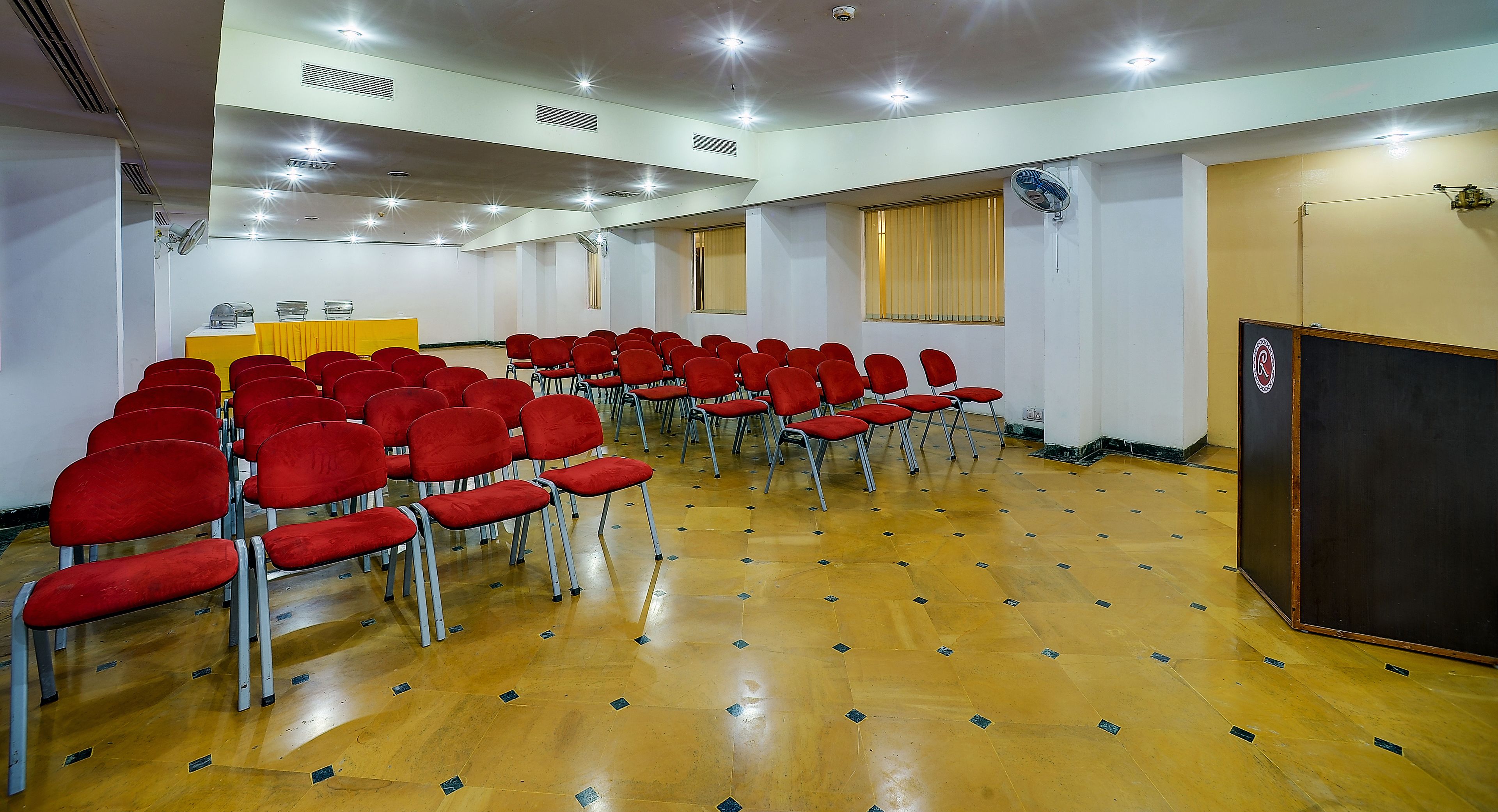 Une salle de conférence avec des rangées de chaises rouges, un pupitre et une grande fenêtre équipée de stores.
