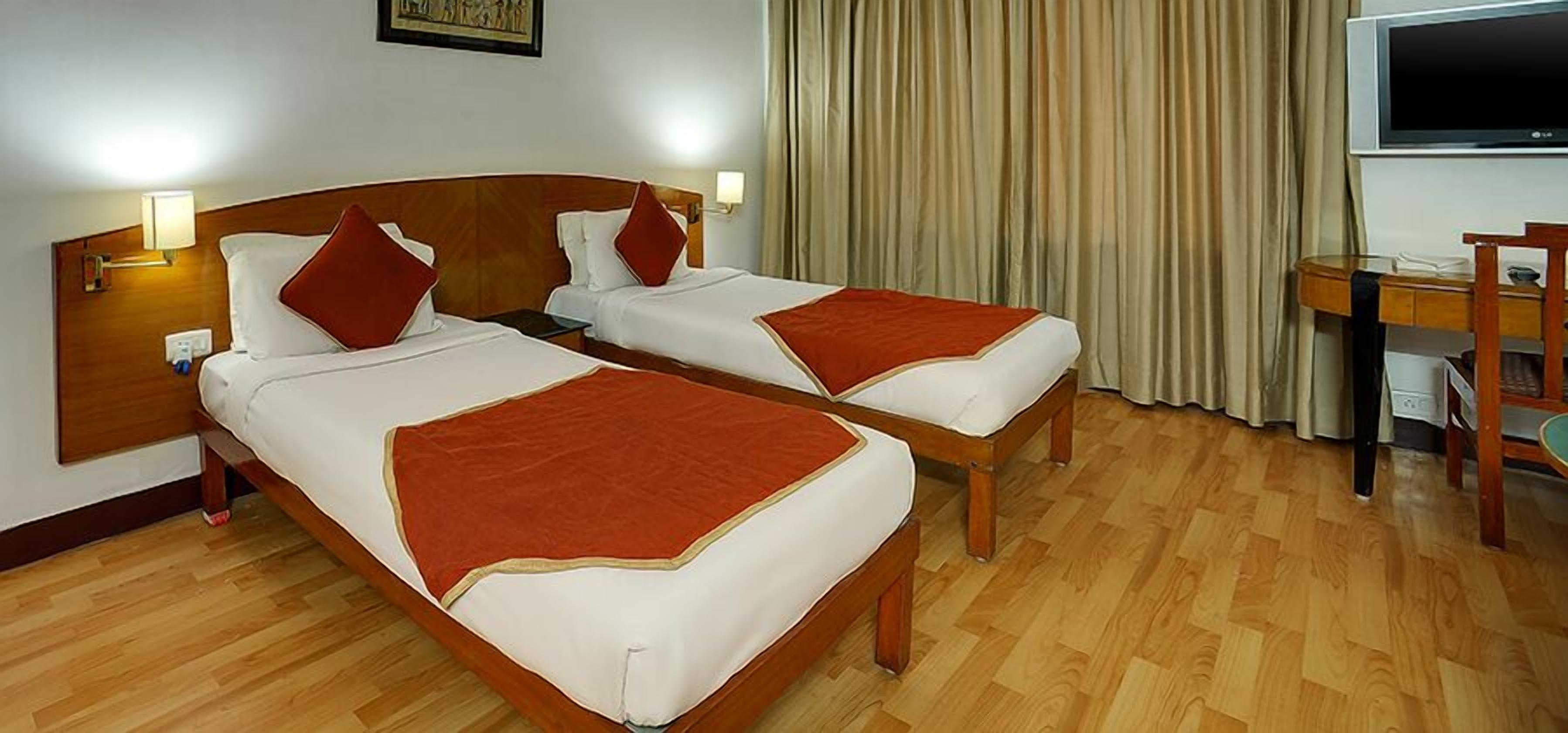 A hotel room with two single beds, wooden headboards, a desk with a chair, and a large window with curtains.