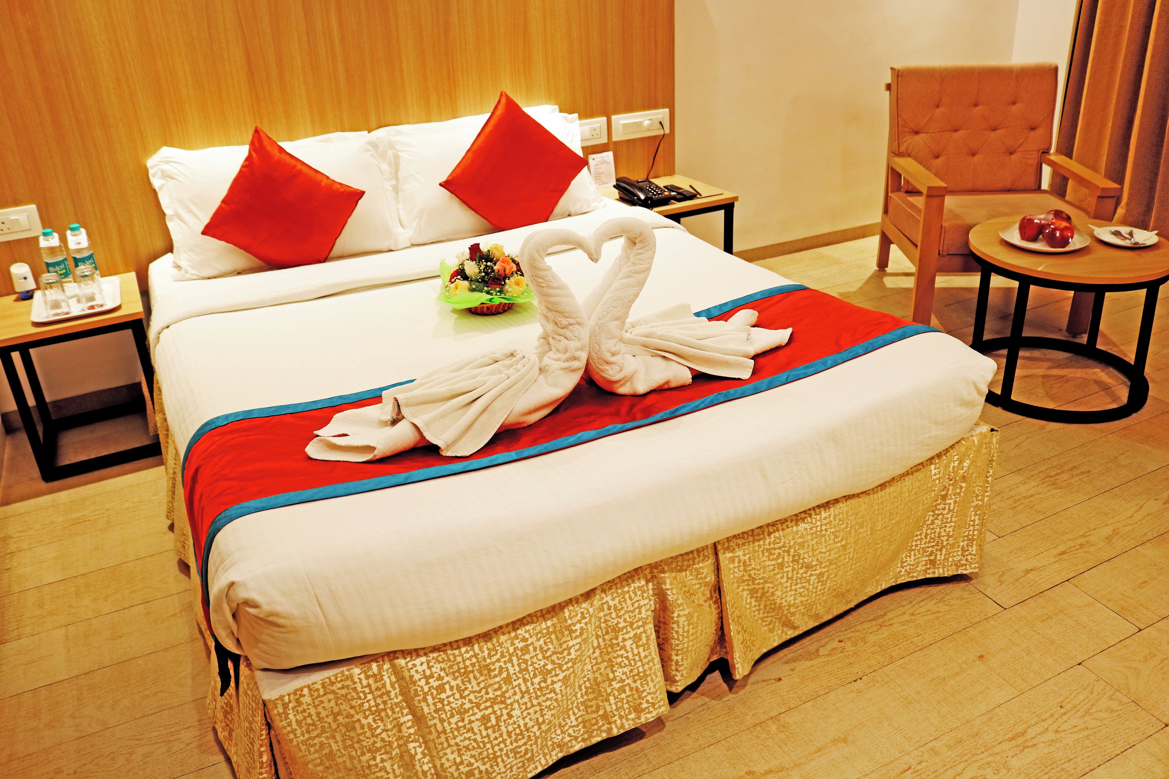 Habitación de hotel con una cama grande, un cabecero de madera, una mesita de noche con un teléfono y botellas de agua, una silla y una pequeña mesa con fruta.