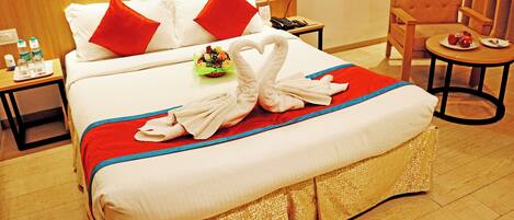 Habitación de hotel con una cama grande, un cabecero de madera, una mesita de noche con un teléfono y botellas de agua, una silla y una pequeña mesa con fruta.