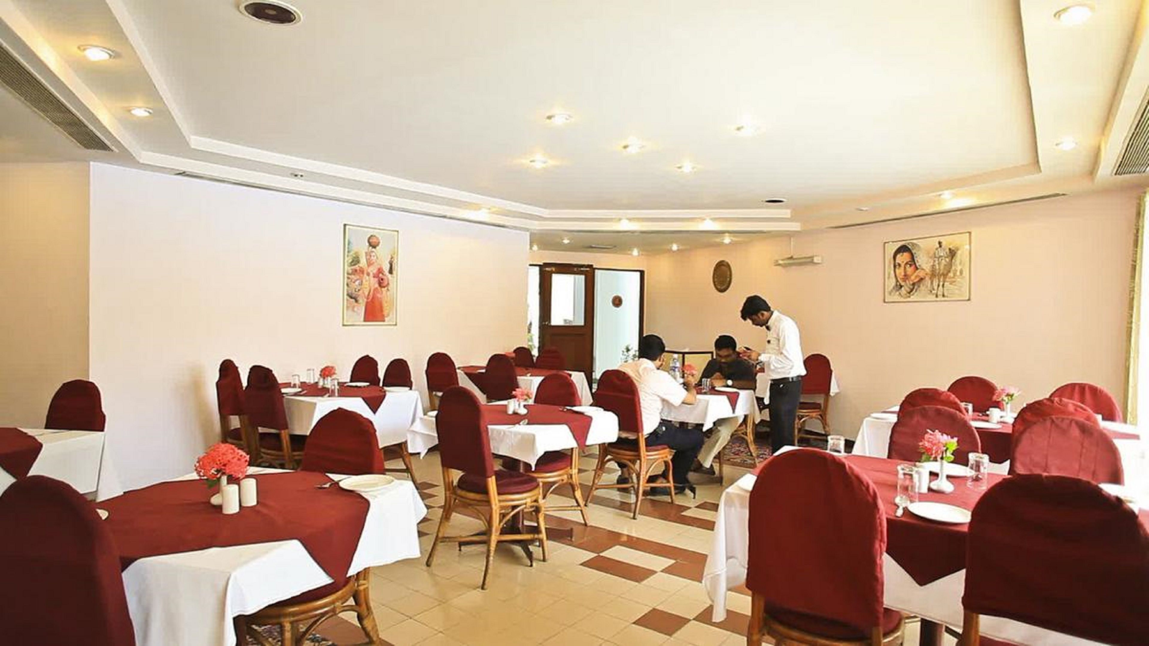 Un espace repas comprenant plusieurs tables et chaises, chacune recouverte d’une nappe blanche et de housses de chaise rouges. On y trouve des luminaires au plafond et des tableaux accrochés aux murs.