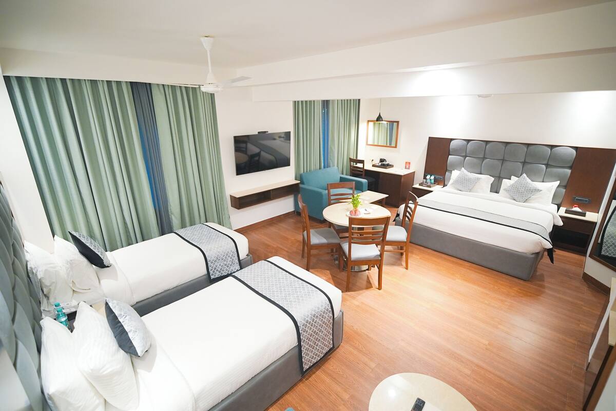 A hotel room with a bed, a sofa, a dining table with chairs, a TV, and a small table with a plant.