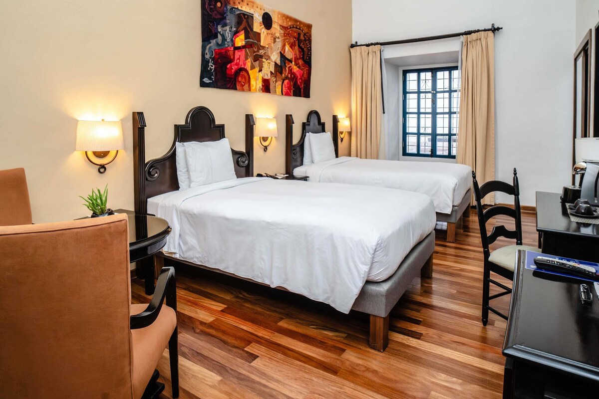 A hotel room with two beds, a desk, a chair, a lamp, a painting on the wall, and a window with curtains.