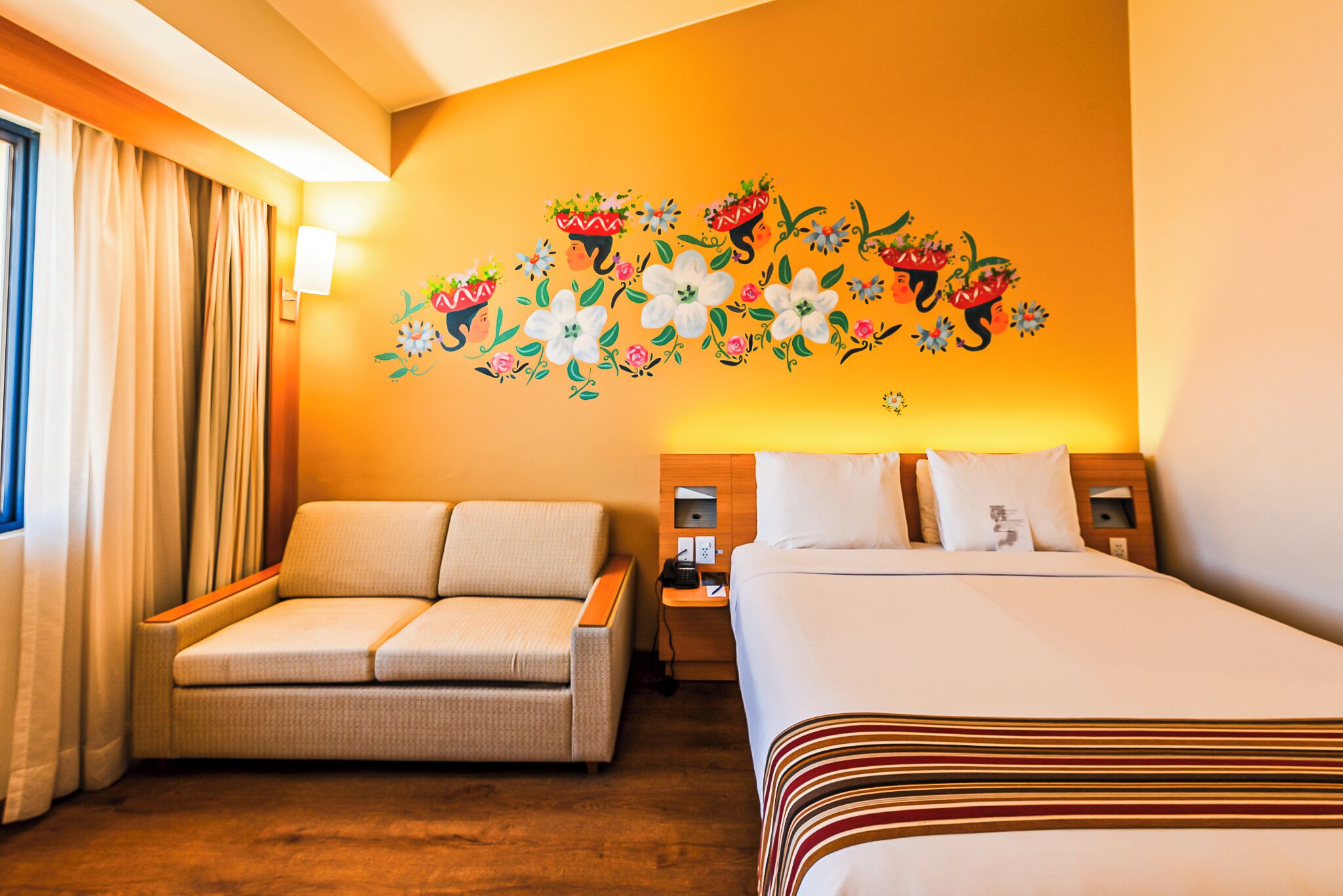 Quarto de hotel com sofá, cama, mesa de cabeceira e parede decorada com arte floral.
