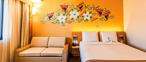 Quarto de hotel com sofá, cama, mesa de cabeceira e parede decorada com arte floral.