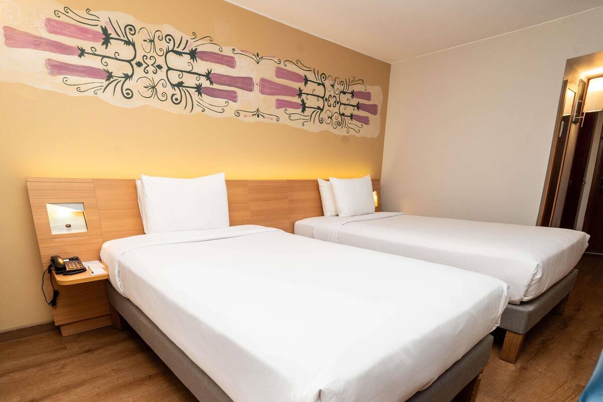 A hotel room with two beds, a wooden headboard, a nightstand with a phone, and a decorative wall piece.