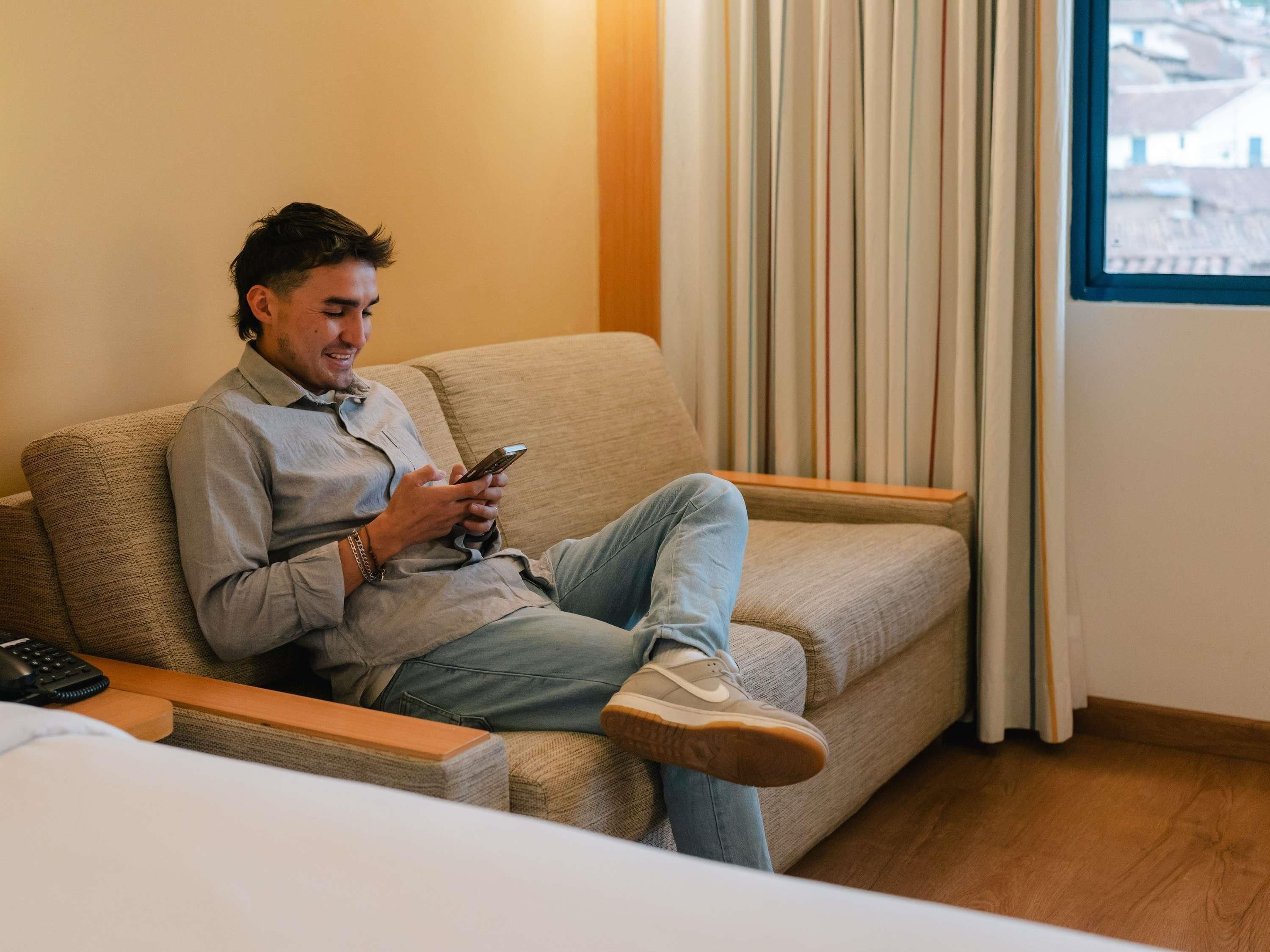 Una persona sentada en un sofá en una habitación con una ventana, un control remoto y un teléfono sobre una mesita.