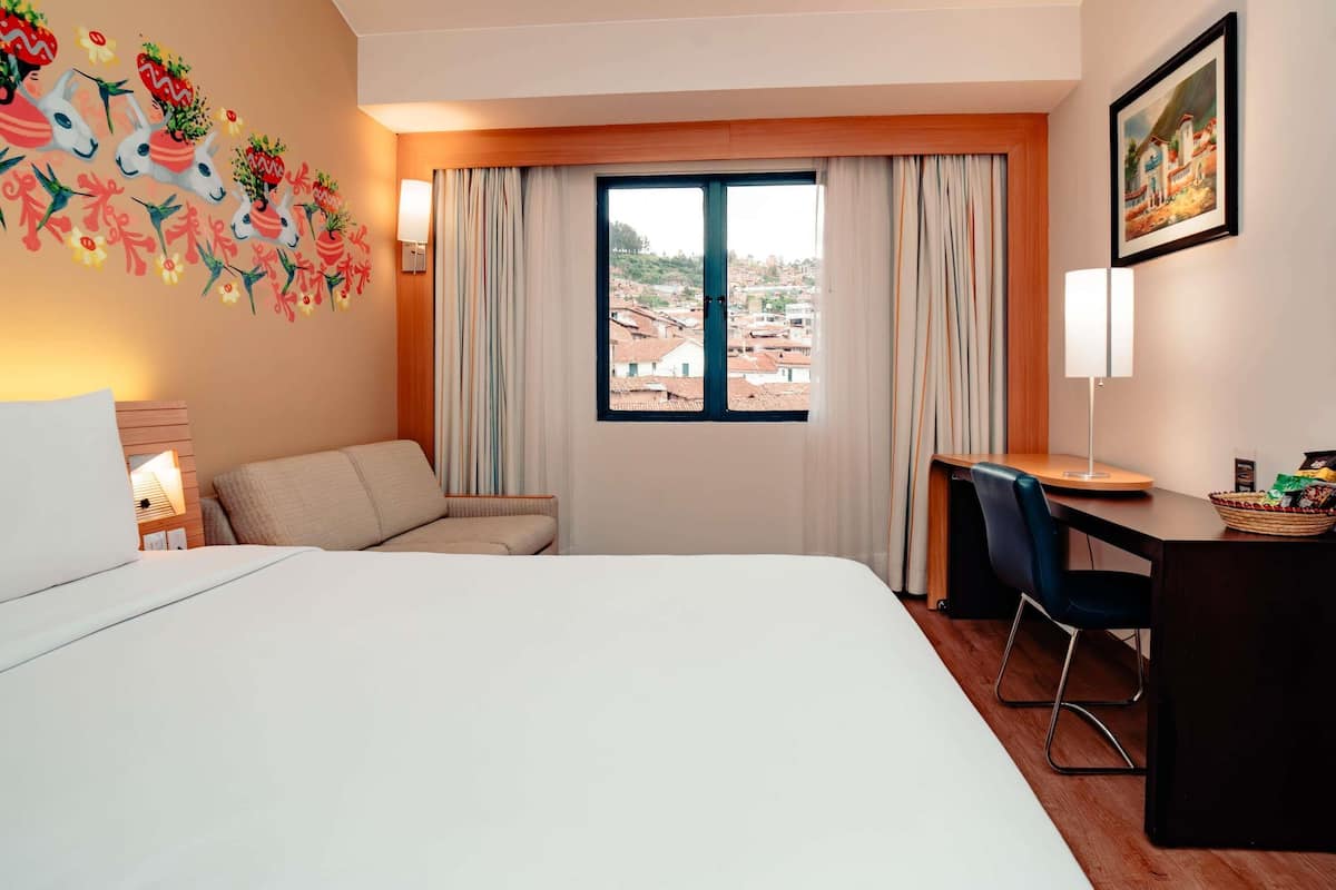 A hotel room with a bed, a desk, a chair, a window with curtains, a wall decoration, and a lamp.