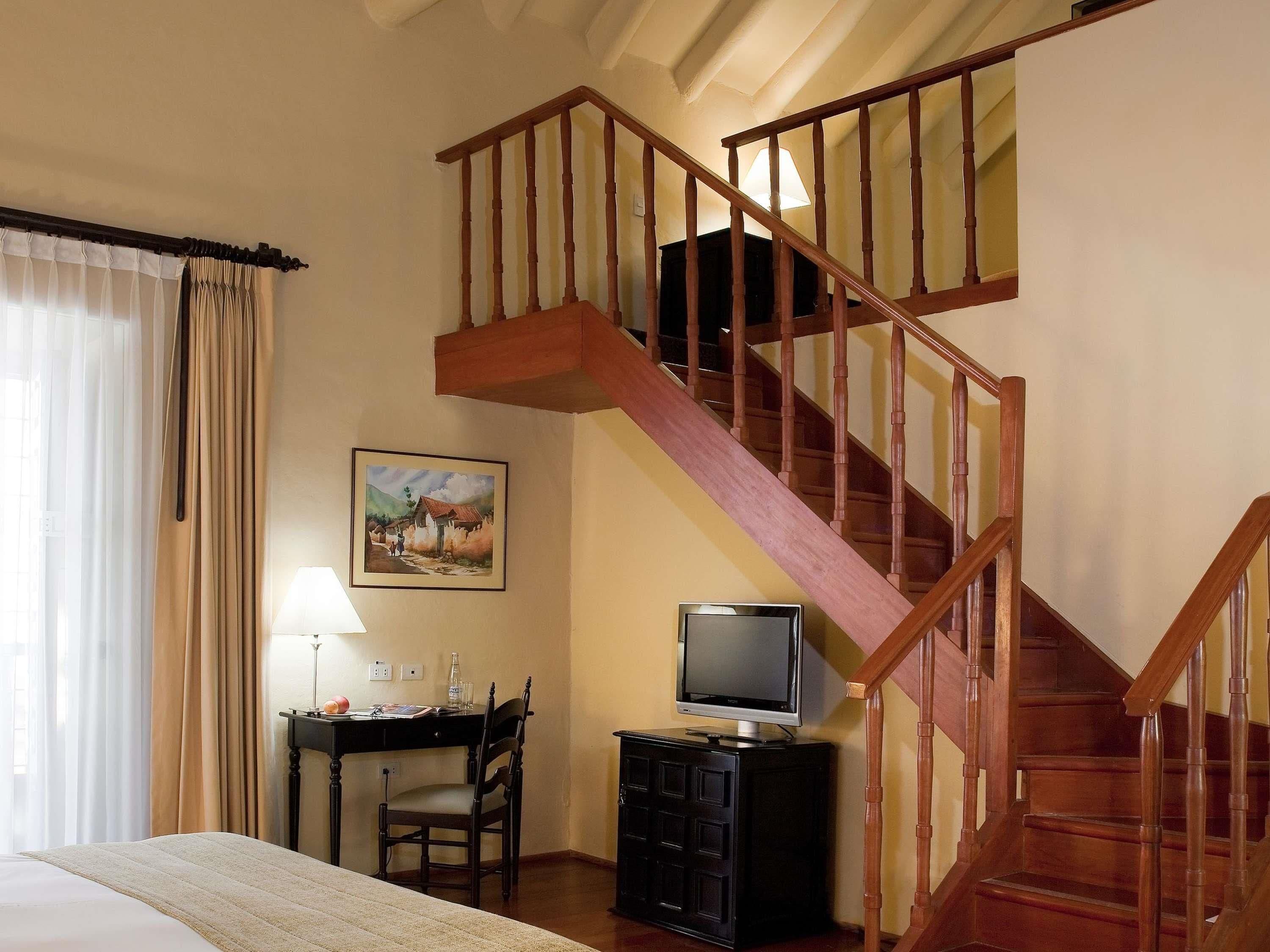 A bedroom with a wooden staircase, a desk with a chair, a television, and a painting on the wall.