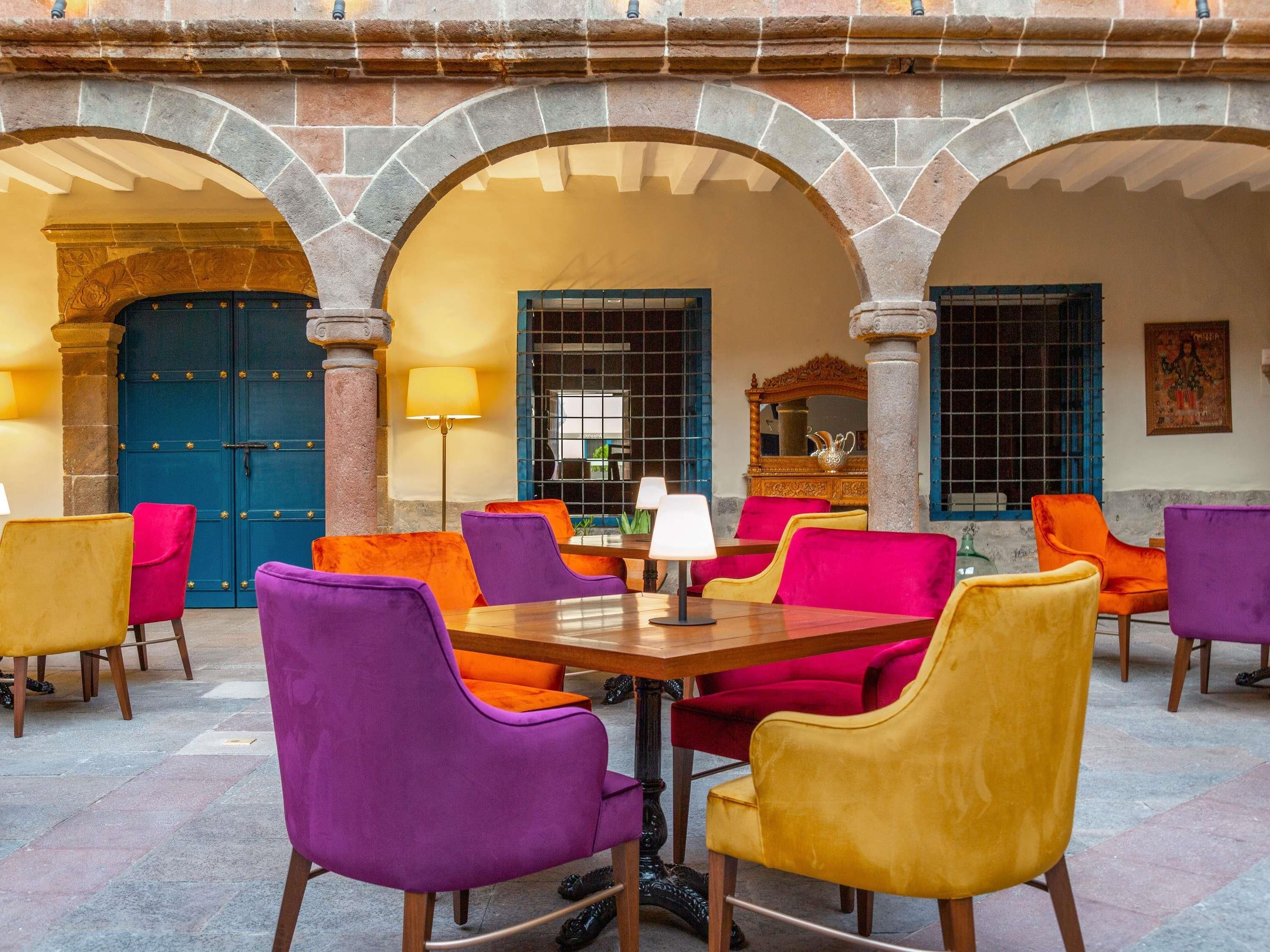 Uma área de refeições com uma mesa central rodeada por cadeiras coloridas, arcos de pedra e uma porta azul.