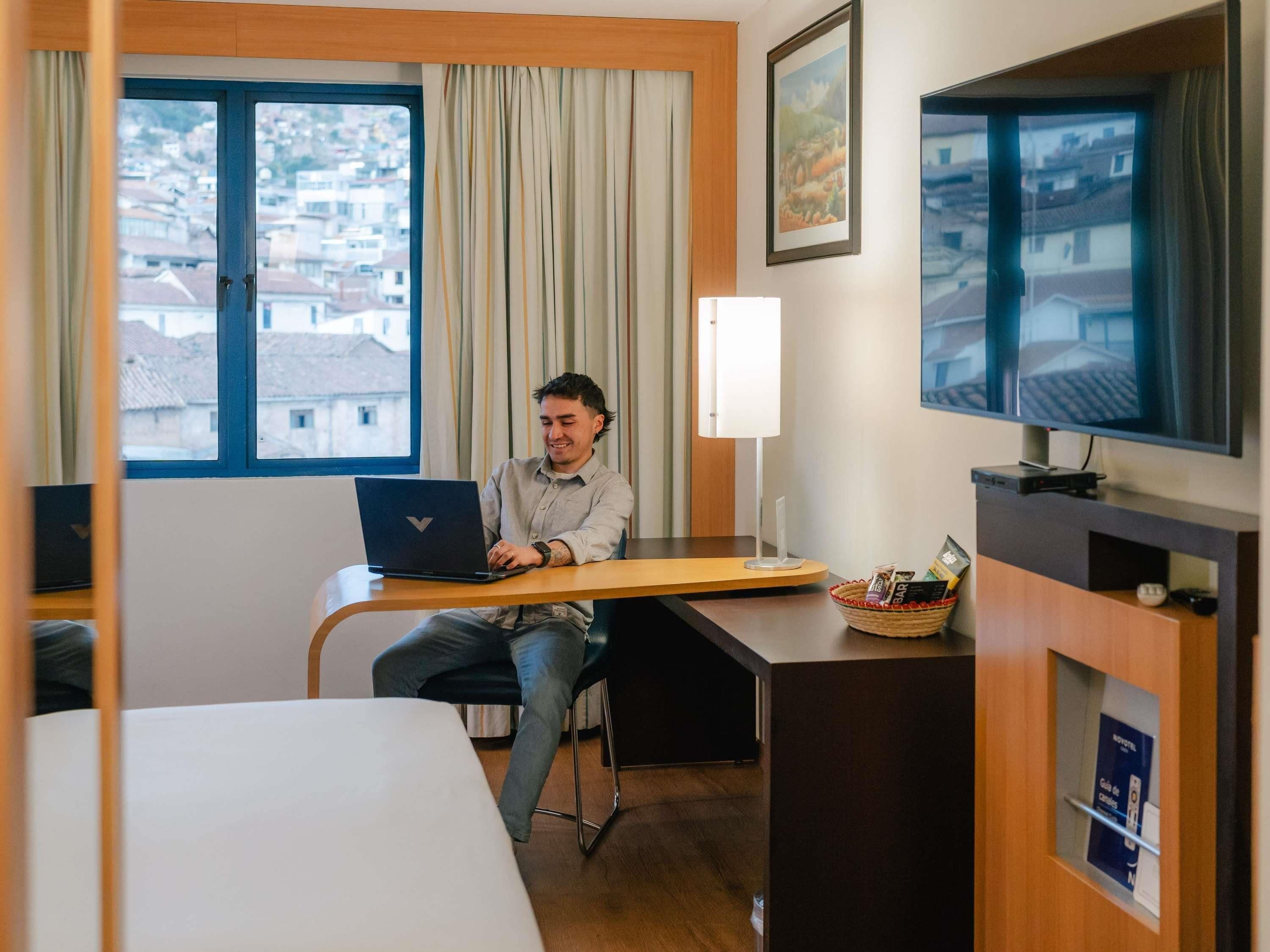 A hotel room with a bed, a desk with a laptop, a TV, and a window with curtains.