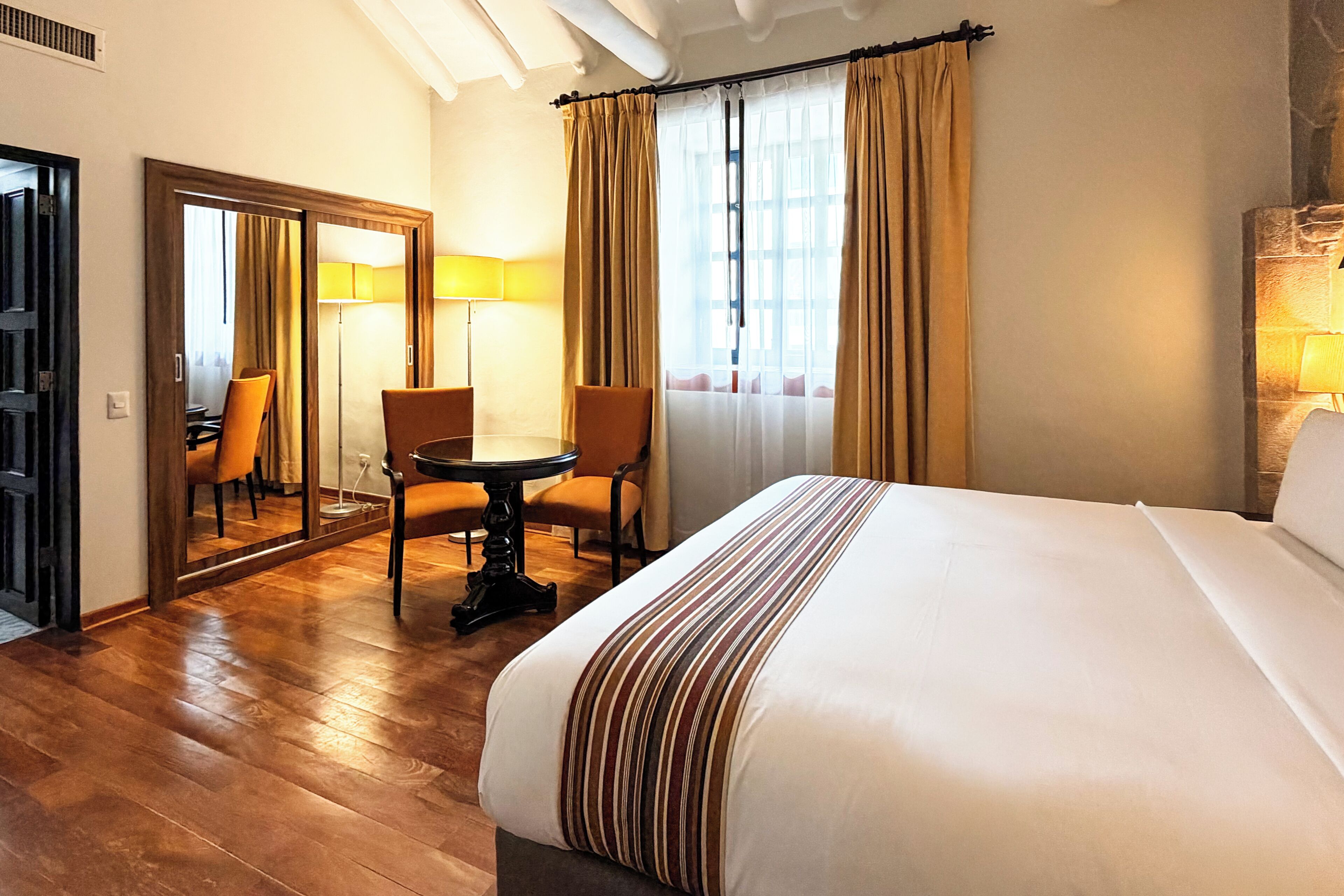 A hotel room with a large bed, two chairs, a small table, and a mirror.