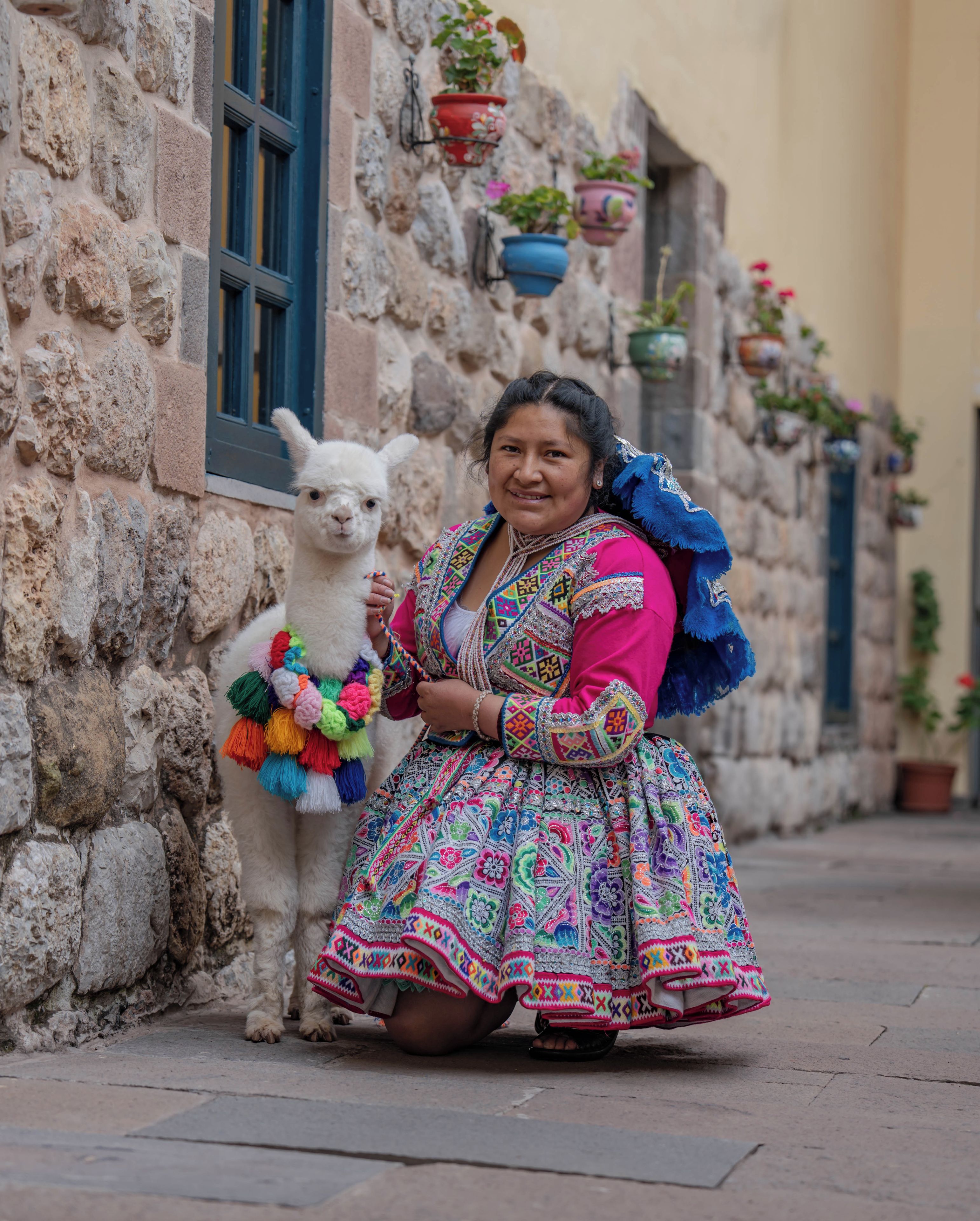 A person in traditional attire sitting next to a llama on a stone-paved street.