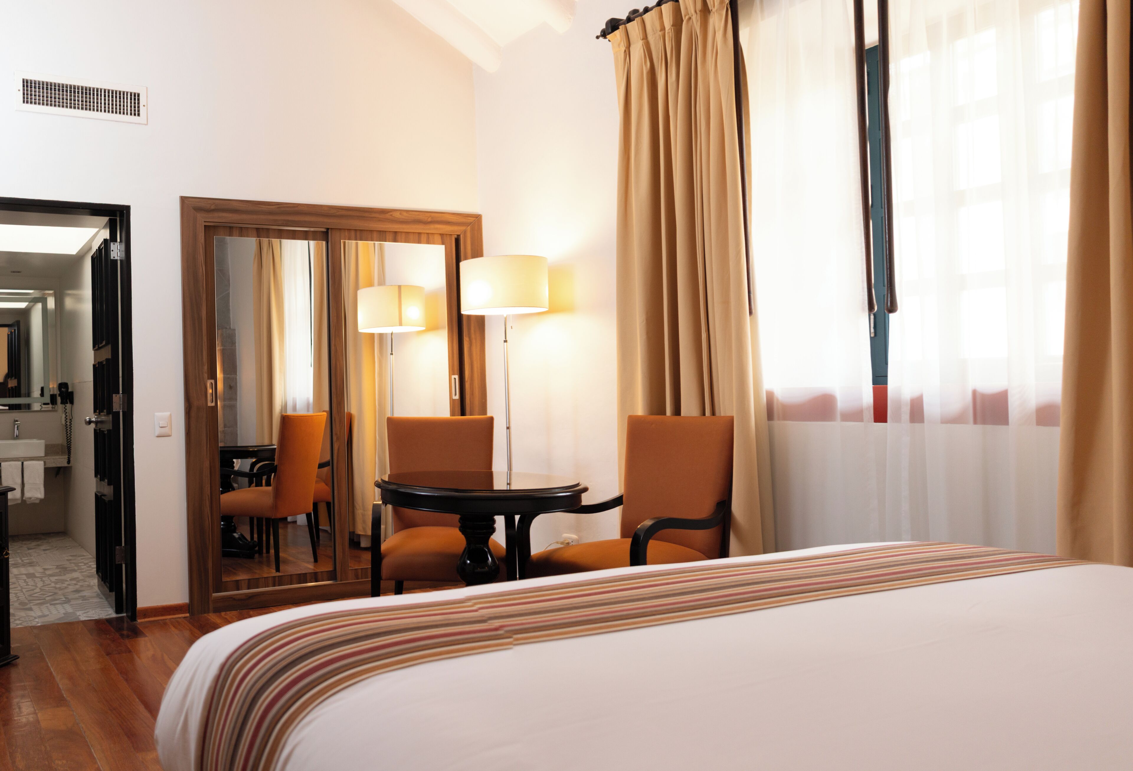 A hotel room with a bed, two chairs, a table, a mirror, and a window with curtains.