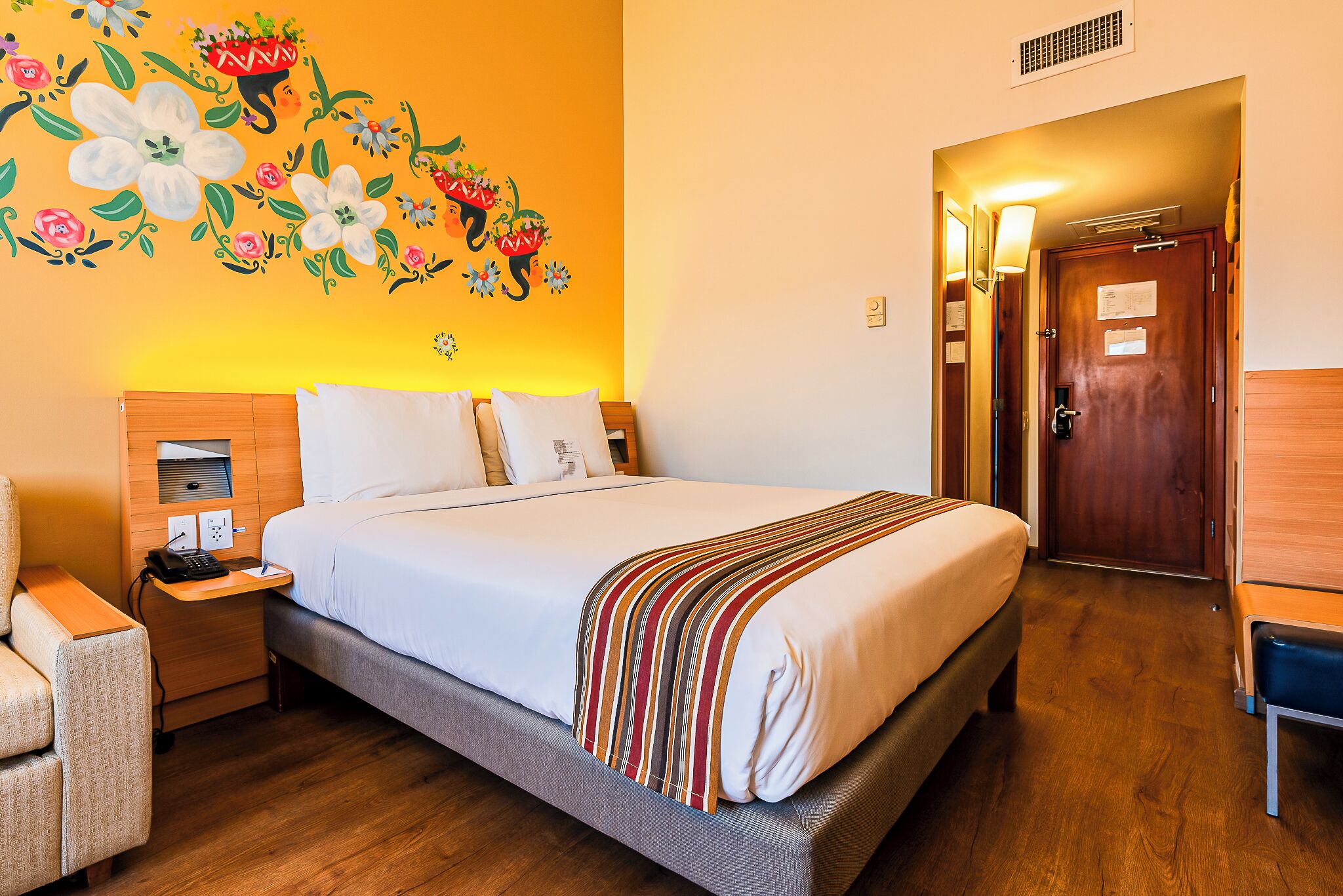 Habitación de hotel con cama, mesita de noche, lámpara y un cuadro floral en la pared.