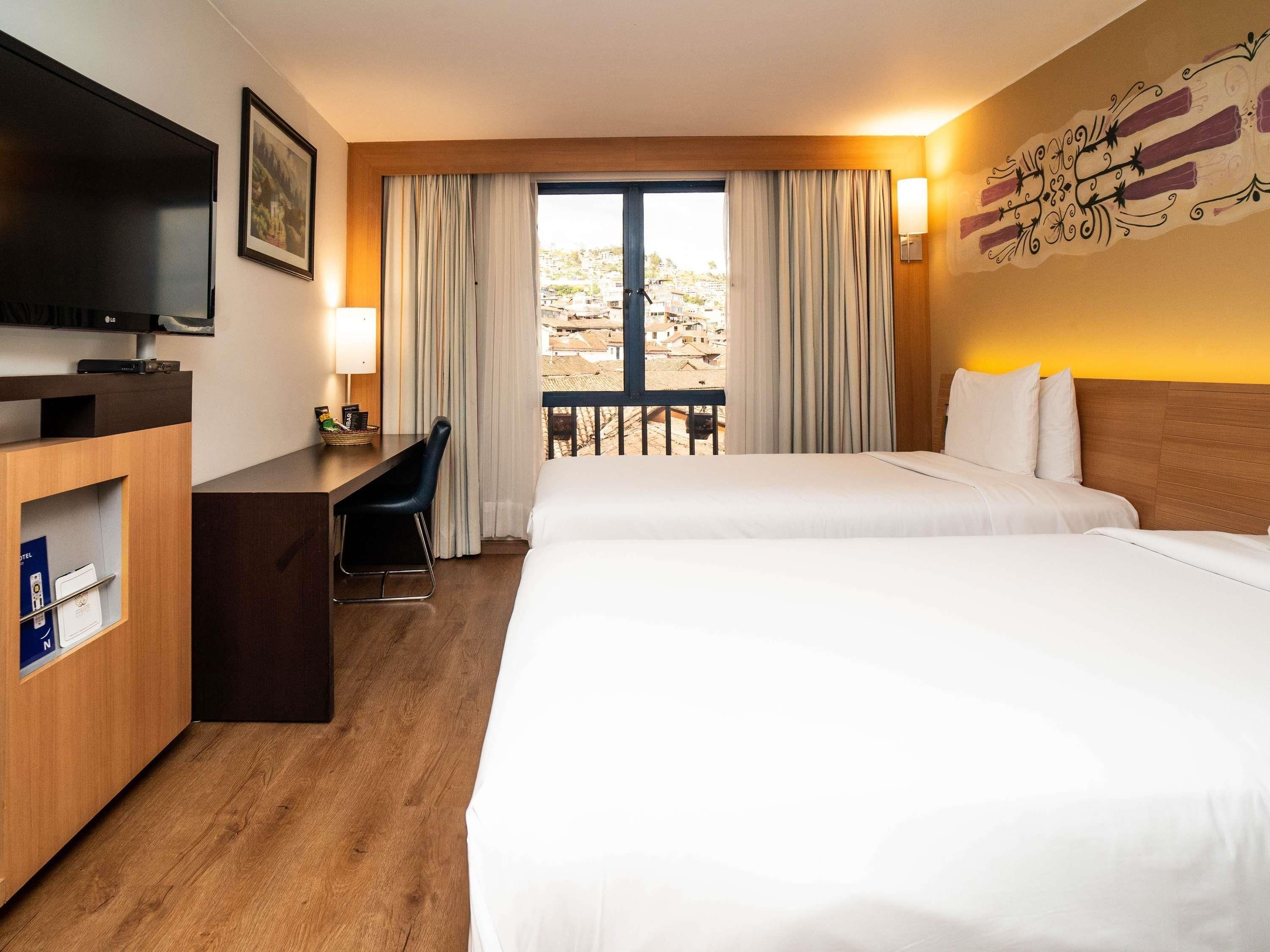 Una habitación de hotel con dos camas, un escritorio con silla, televisión y balcón con cortinas.