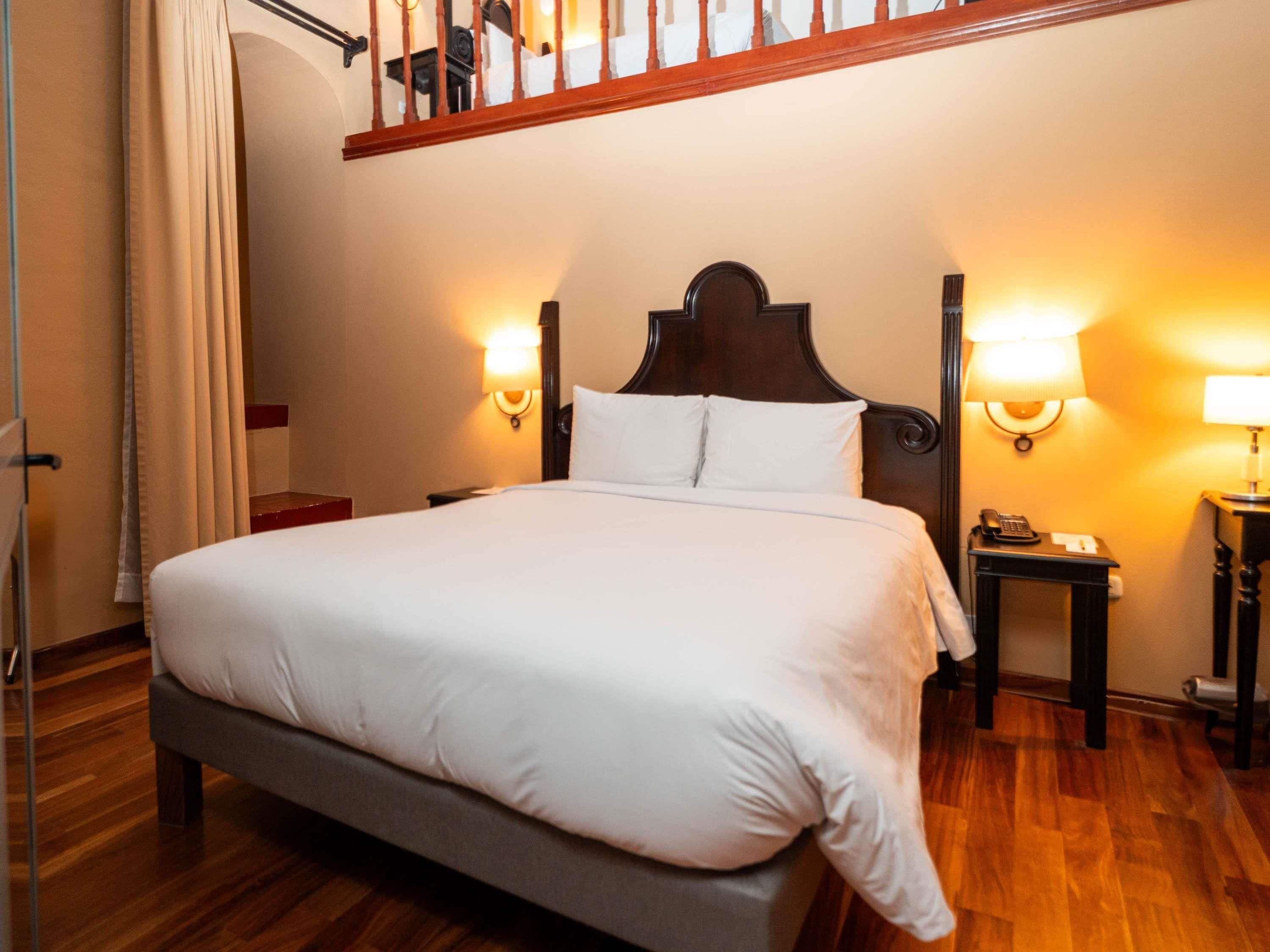 Quarto de hotel com uma cama grande, piso de madeira, uma escada e duas luminárias de cabeceira.