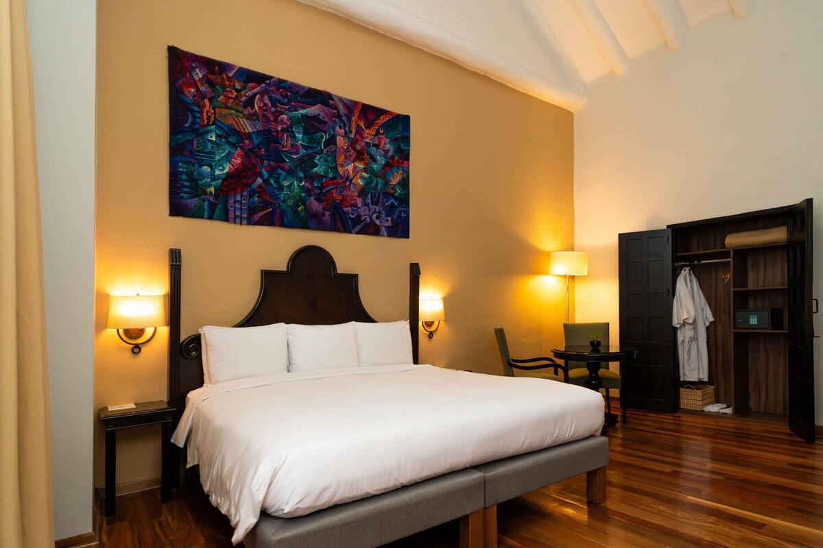 A hotel room with a large bed, two bedside lamps, a wooden floor, a colorful abstract painting on the wall, and a wardrobe with a mirror.