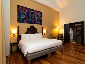 A hotel room with a large bed, two bedside lamps, a wooden floor, a colorful abstract painting on the wall, and a wardrobe with a mirror.