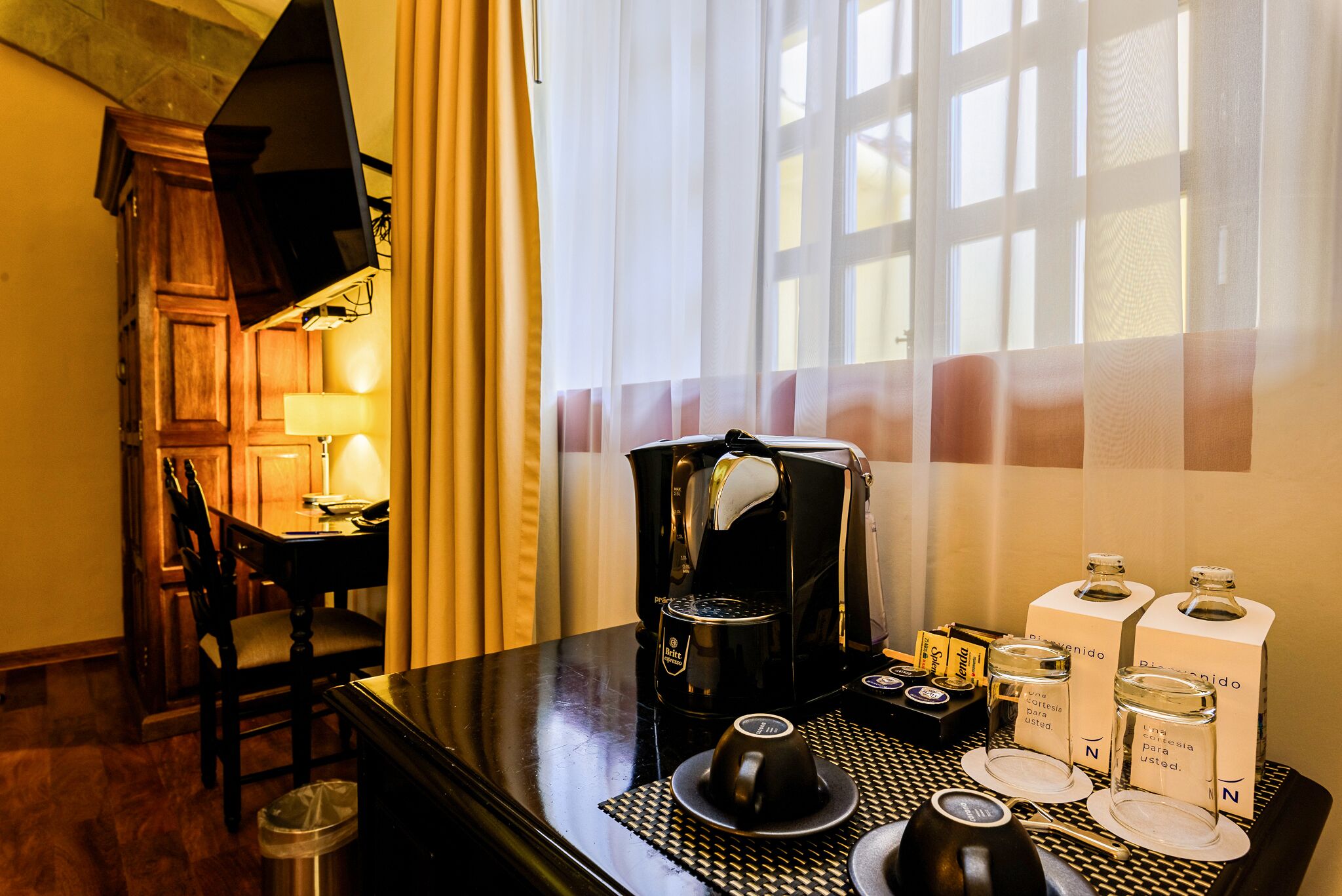 A hotel room with a dining table set for two, a coffee machine, and a window with curtains.