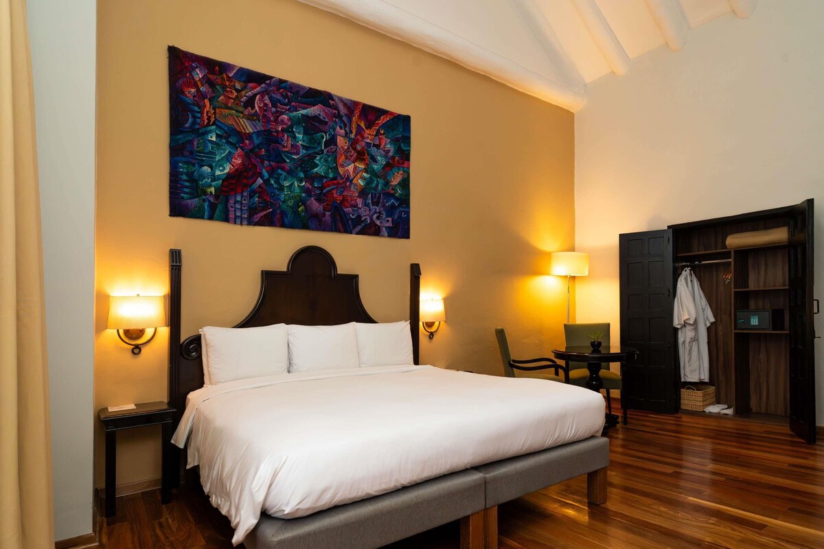A hotel room with a large bed, two bedside lamps, a wooden floor, a colorful abstract painting on the wall, and a wardrobe with a mirror.