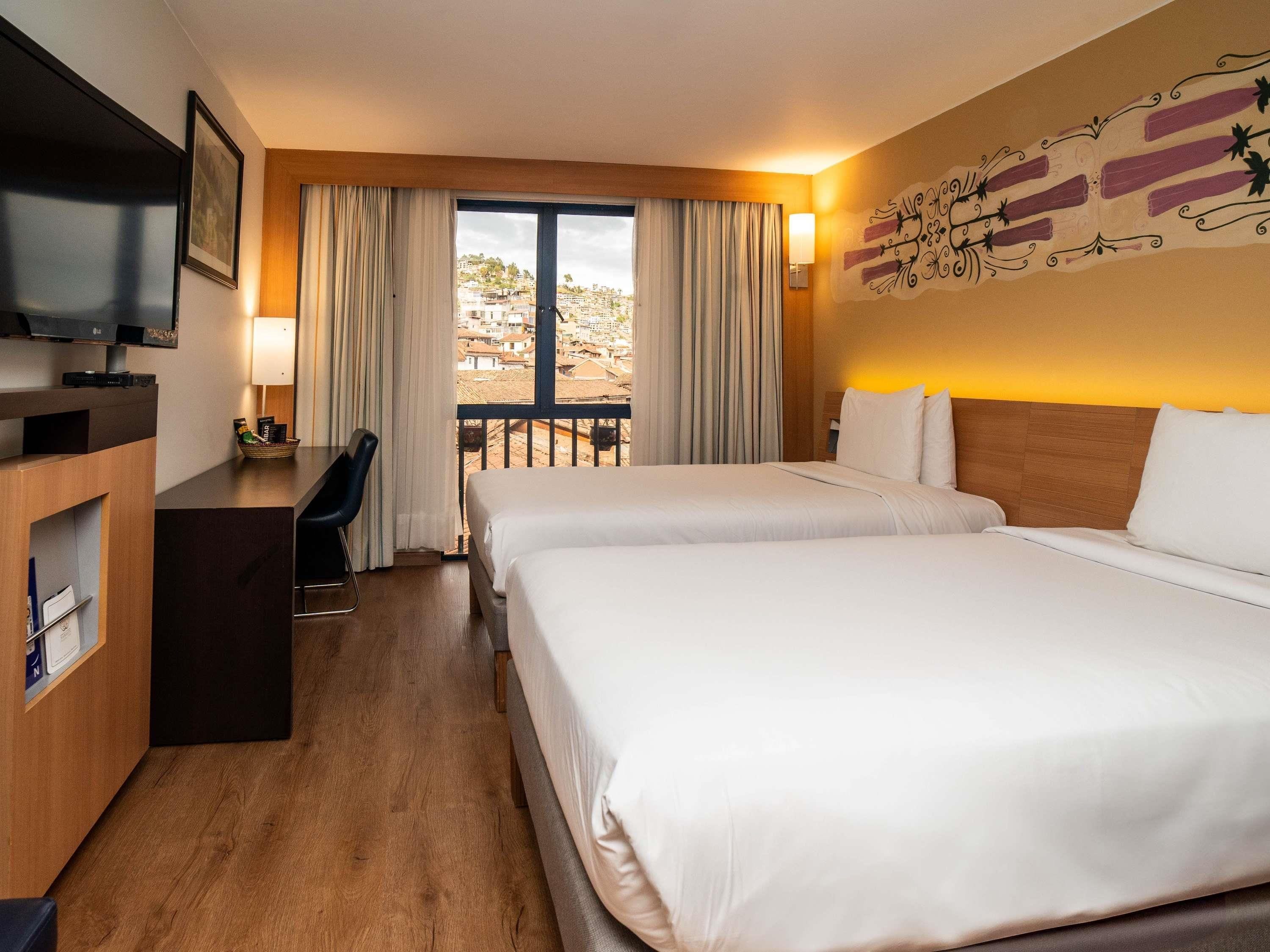 Una habitación de hotel con cama, escritorio, televisión y balcón con cortinas.
