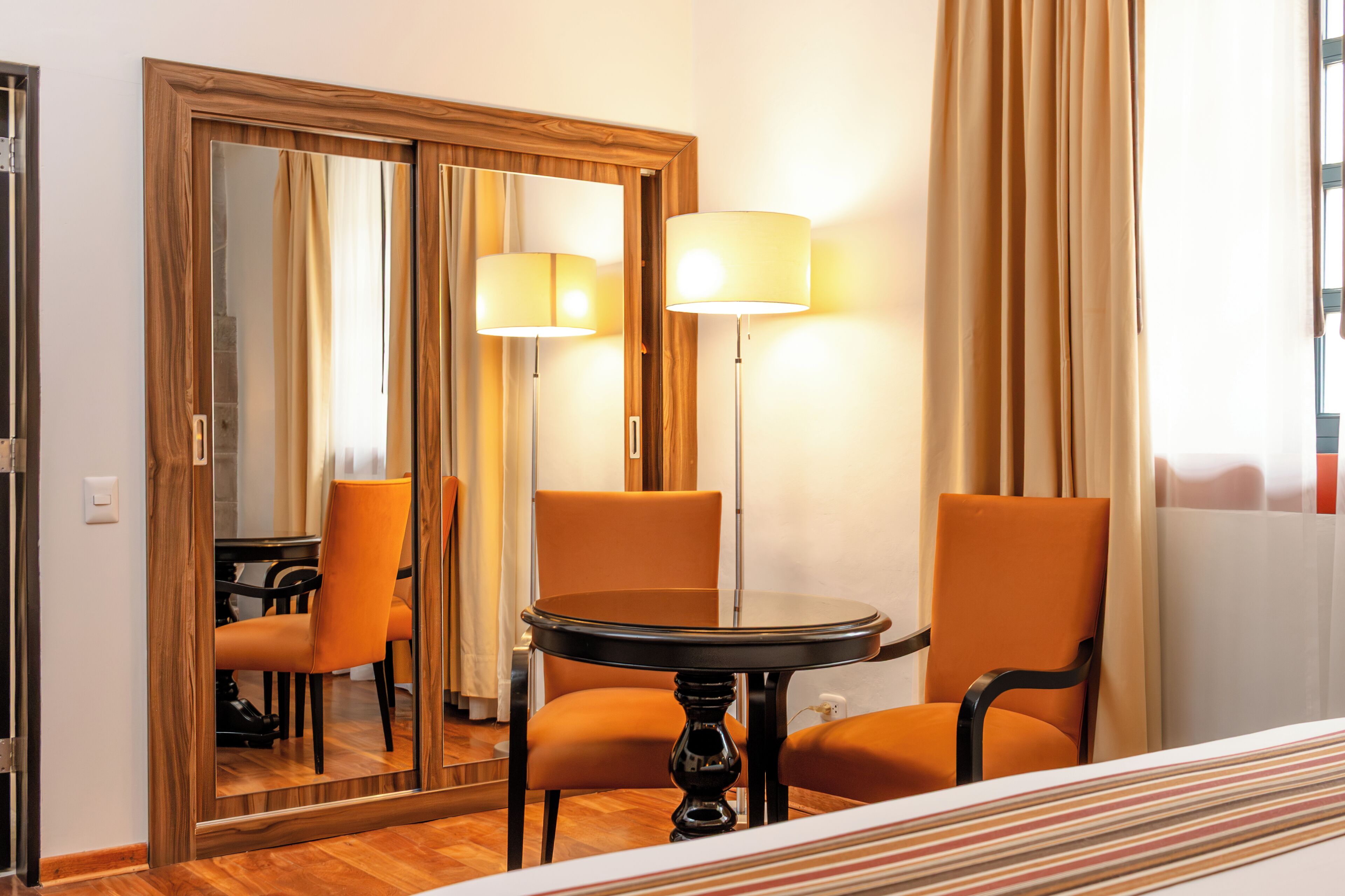 A hotel room with a bed, two chairs, a table, a mirror, and a lamp.