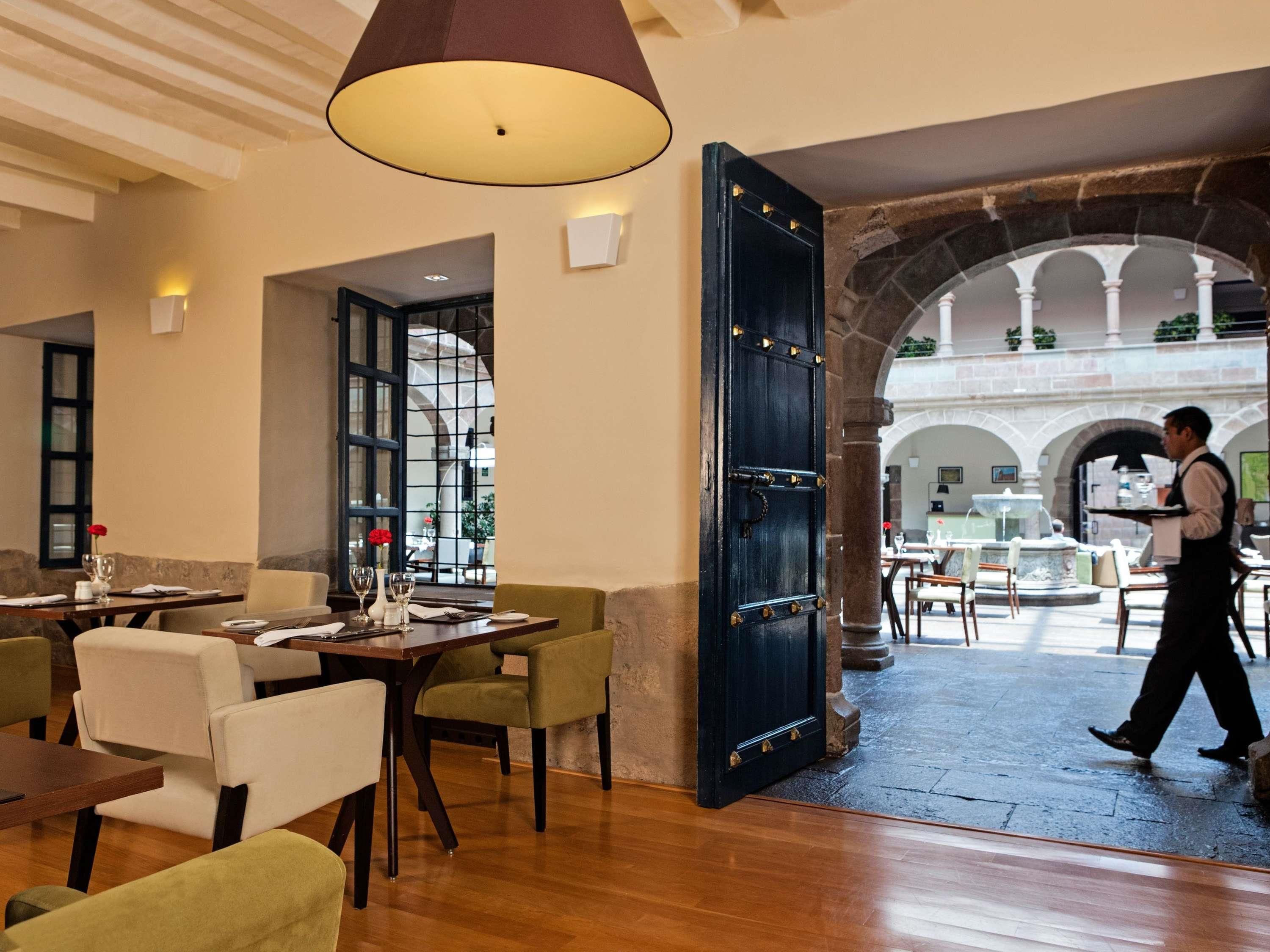 A restaurant with wooden tables and chairs, a waiter carrying a tray, and an open door leading to an outdoor area with stone arches.