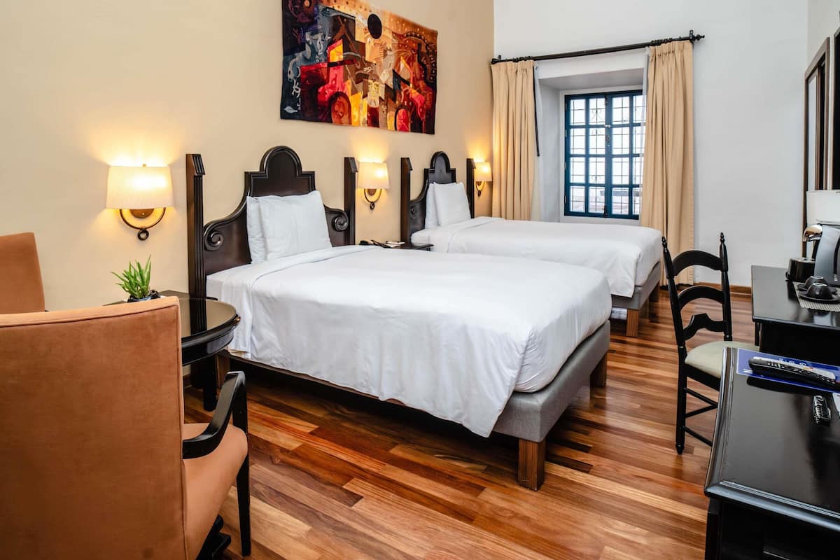 A hotel room with two beds, a desk, a chair, a lamp, a painting on the wall, and a window with curtains.