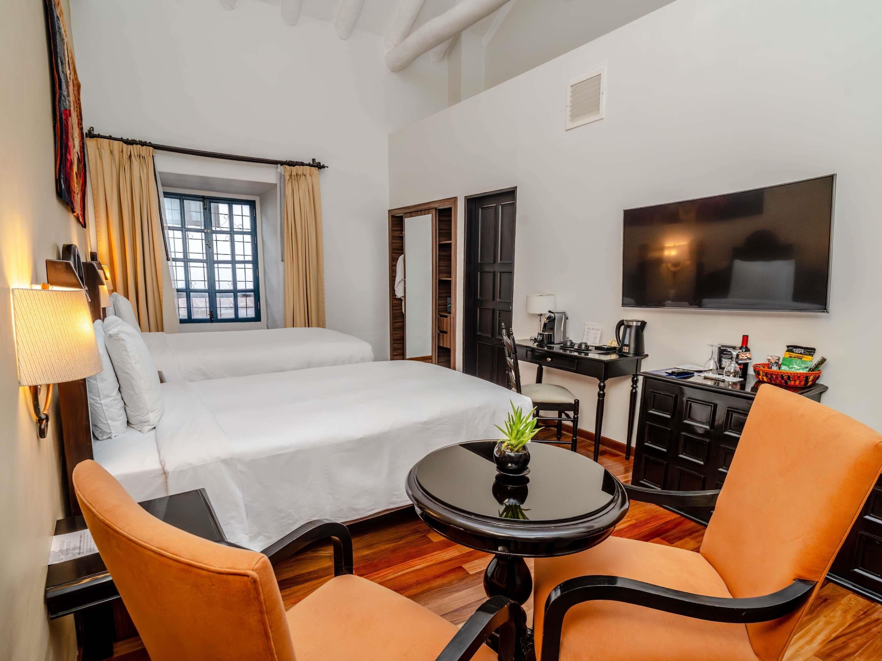 A hotel room with a bed, a desk with a chair, a small table with a plant, and a television on the wall.