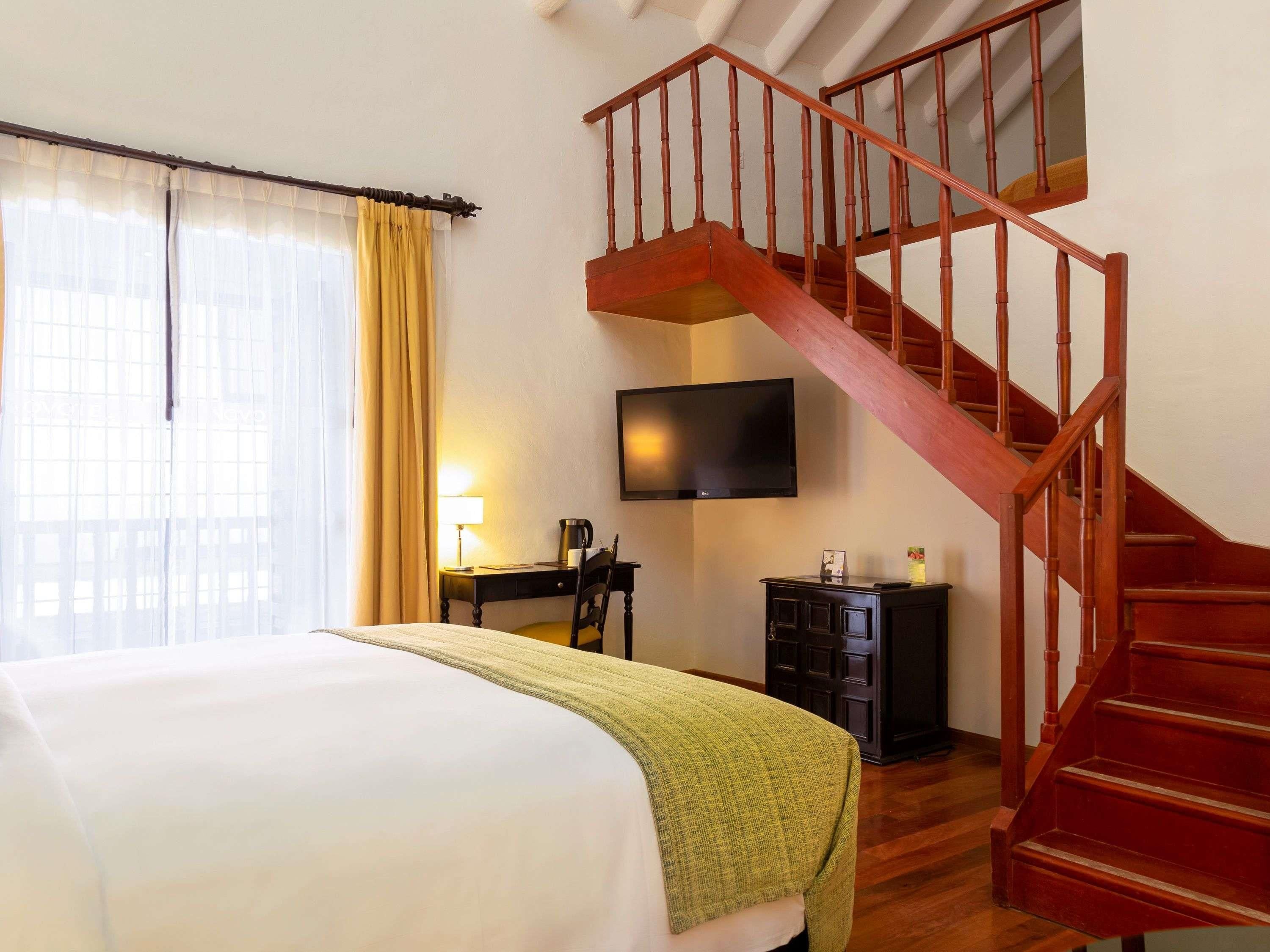 A hotel room with a bed, a desk with a lamp, a television, a staircase, and a wardrobe.