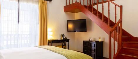 A hotel room with a bed, a desk with a lamp, a television, a staircase, and a wardrobe.
