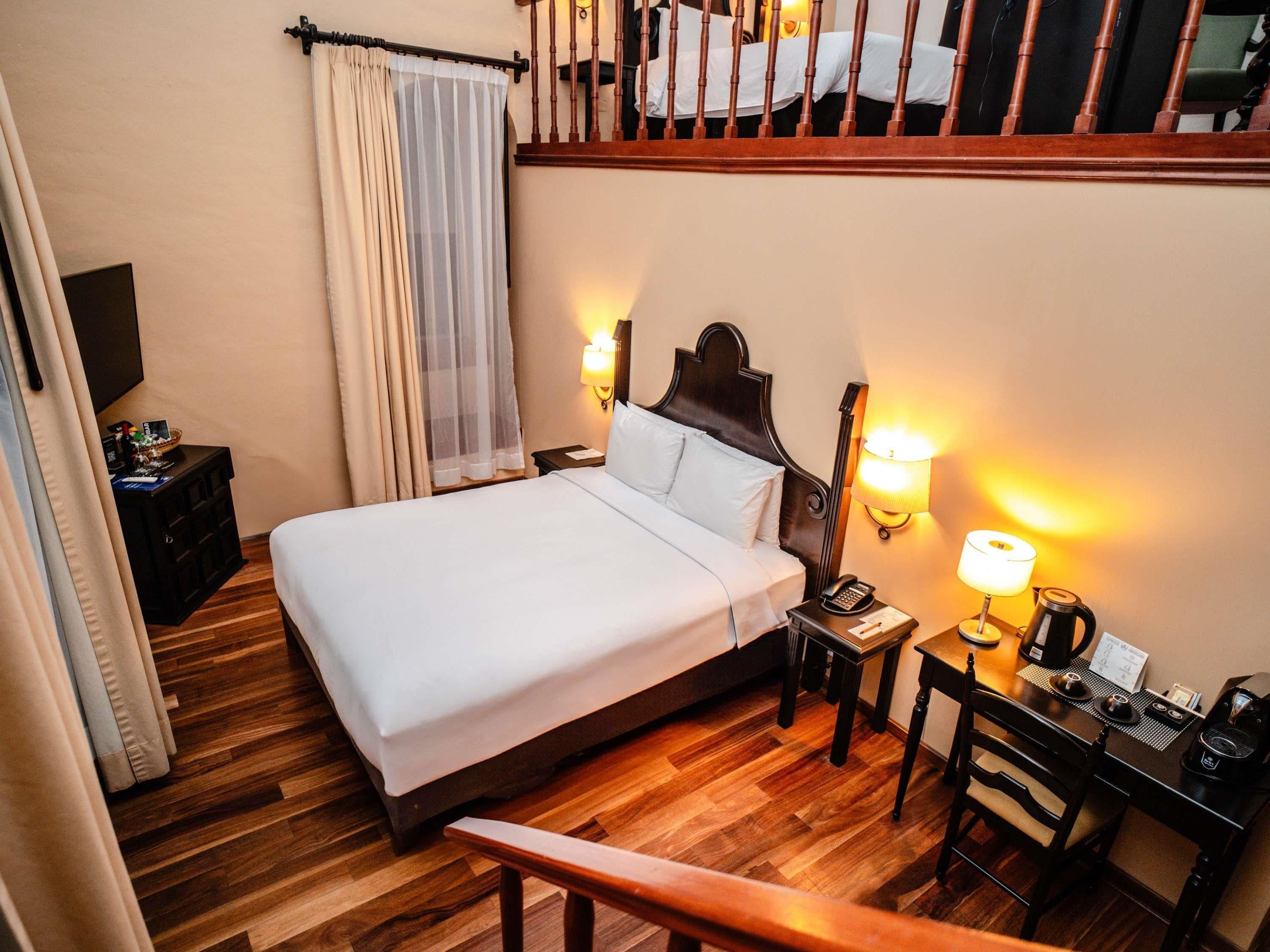 Une chambre d’hôtel avec un lit, des lampes de chevet, un bureau avec un téléphone et une bouilloire, une chaise et un plancher en bois.