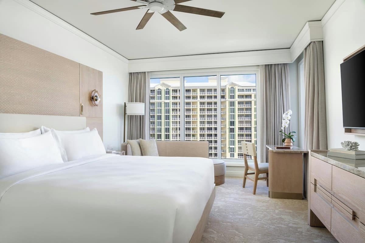 A hotel room with a large bed, a desk, a chair, a TV, and a view of high-rise buildings.