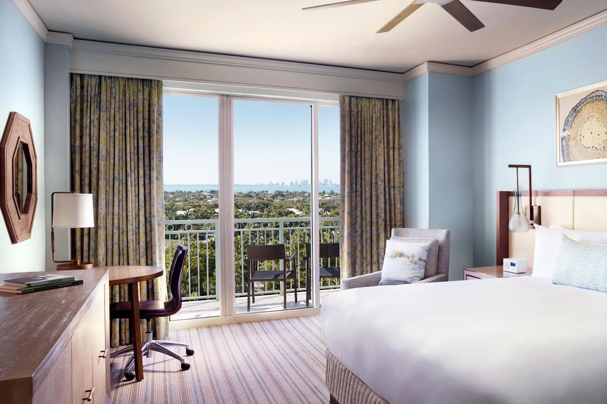 A hotel room with a large bed, a desk with a chair, a nightstand, and a view of the city through large windows.