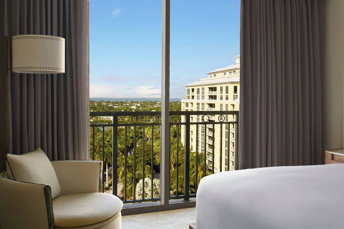 A hotel room with a large window, a bed, a chair, and a lamp.