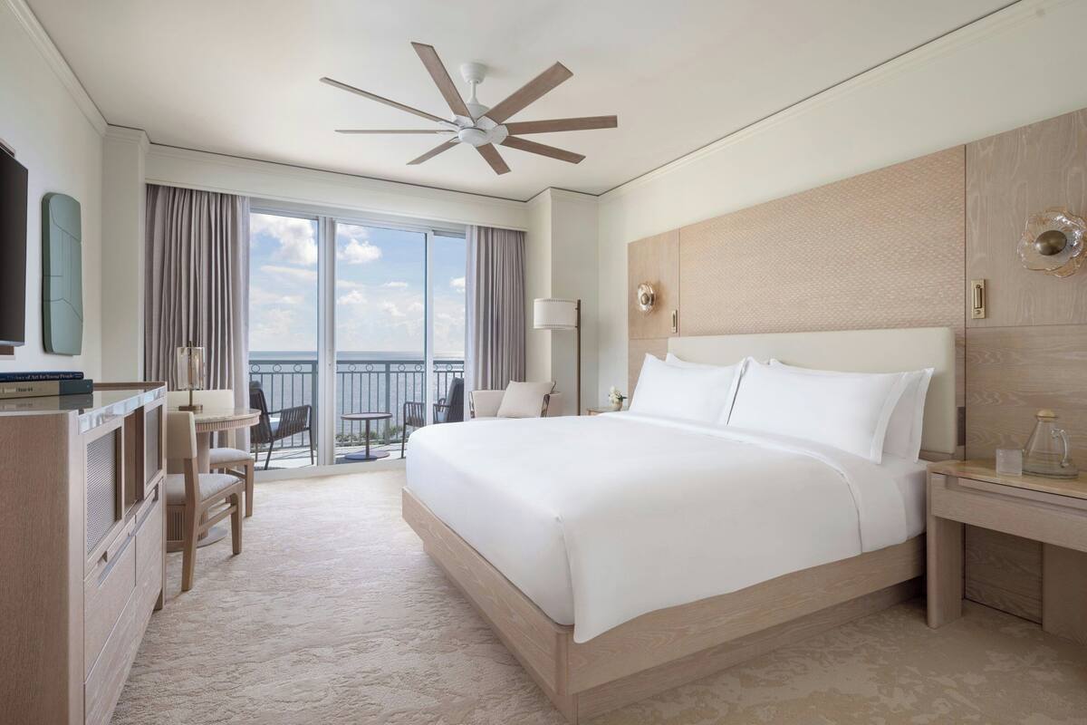 A hotel room with a large bed, a desk, a chair, a ceiling fan, and a view of the outdoors.