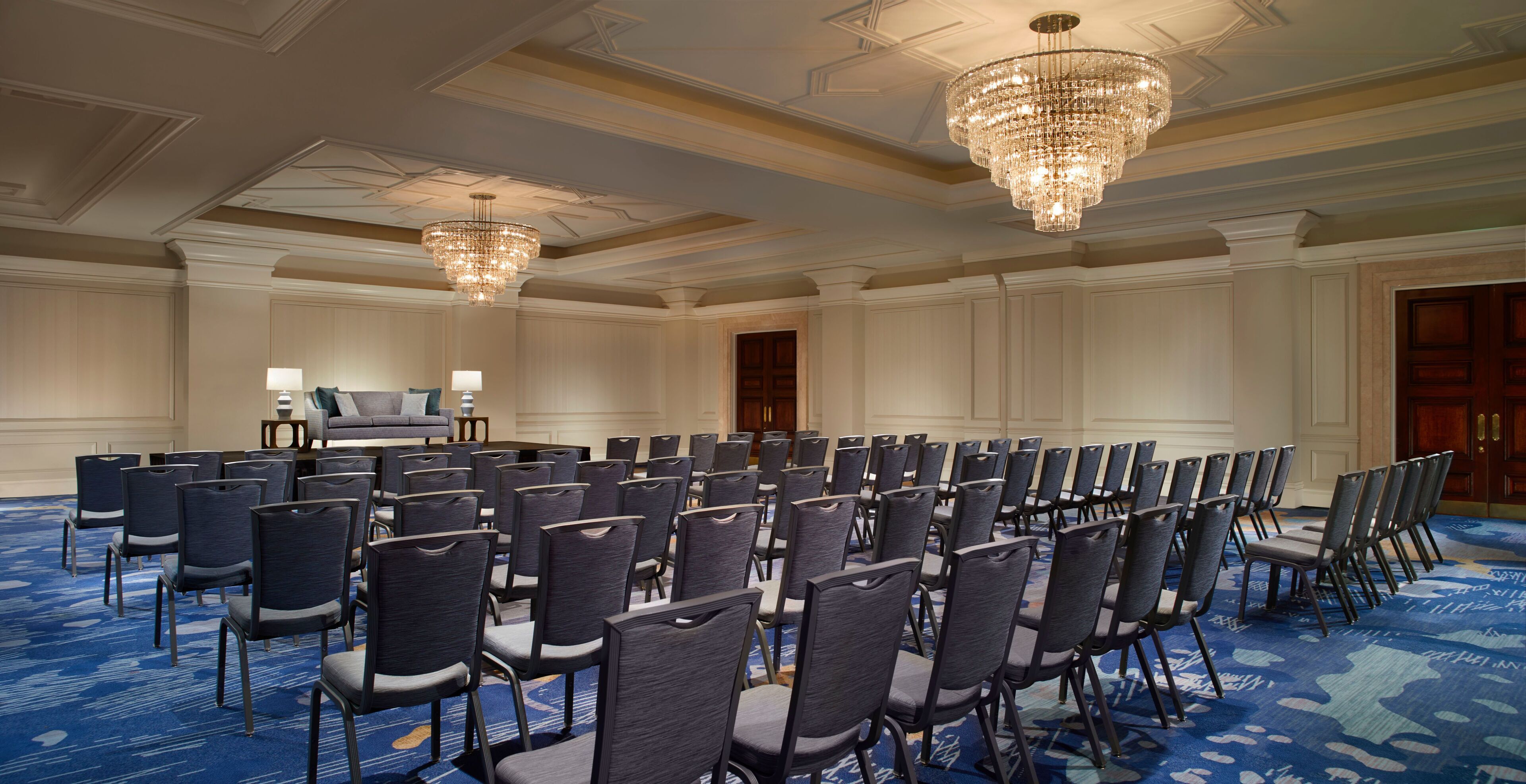 A conference room with rows of chairs, a stage with a podium, and chandeliers.