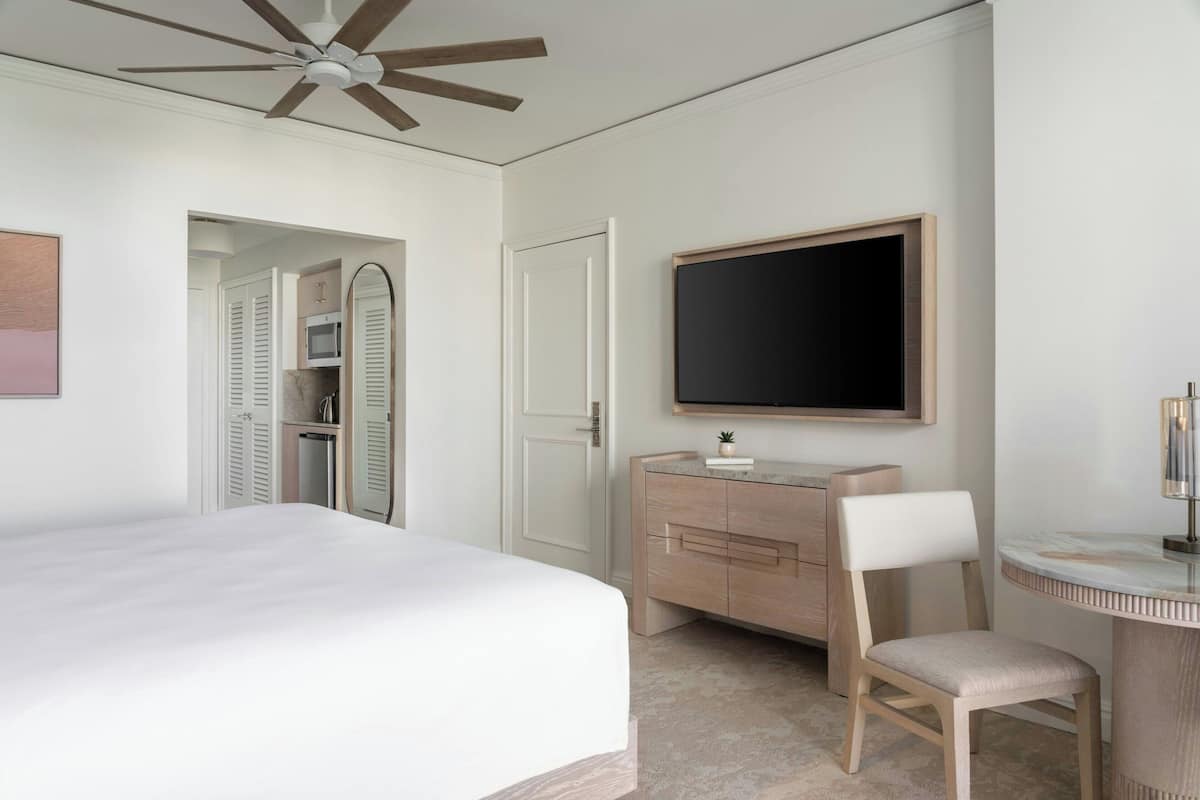 A hotel room with a bed, a vanity with a mirror, a television mounted on the wall, and a ceiling fan.