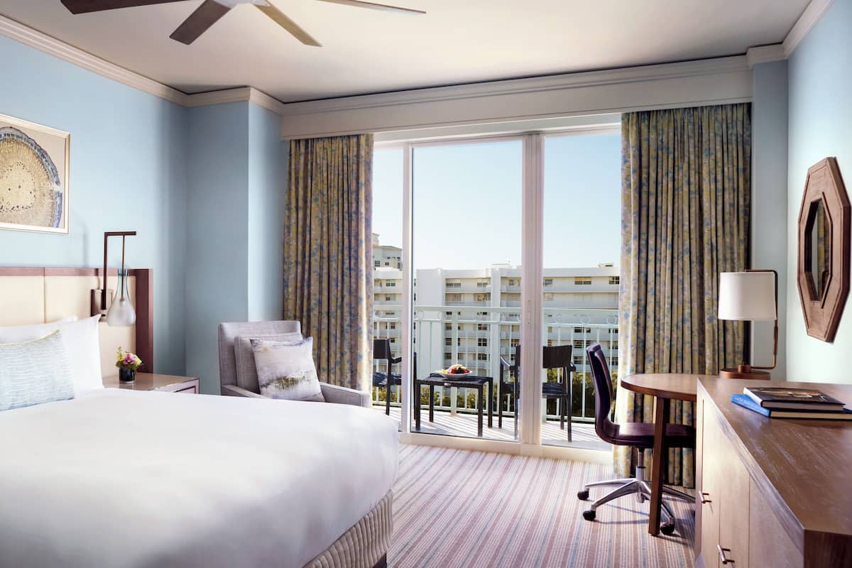 A hotel room with a large bed, a desk, a chair, a balcony with a view of buildings, and two artworks on the wall.